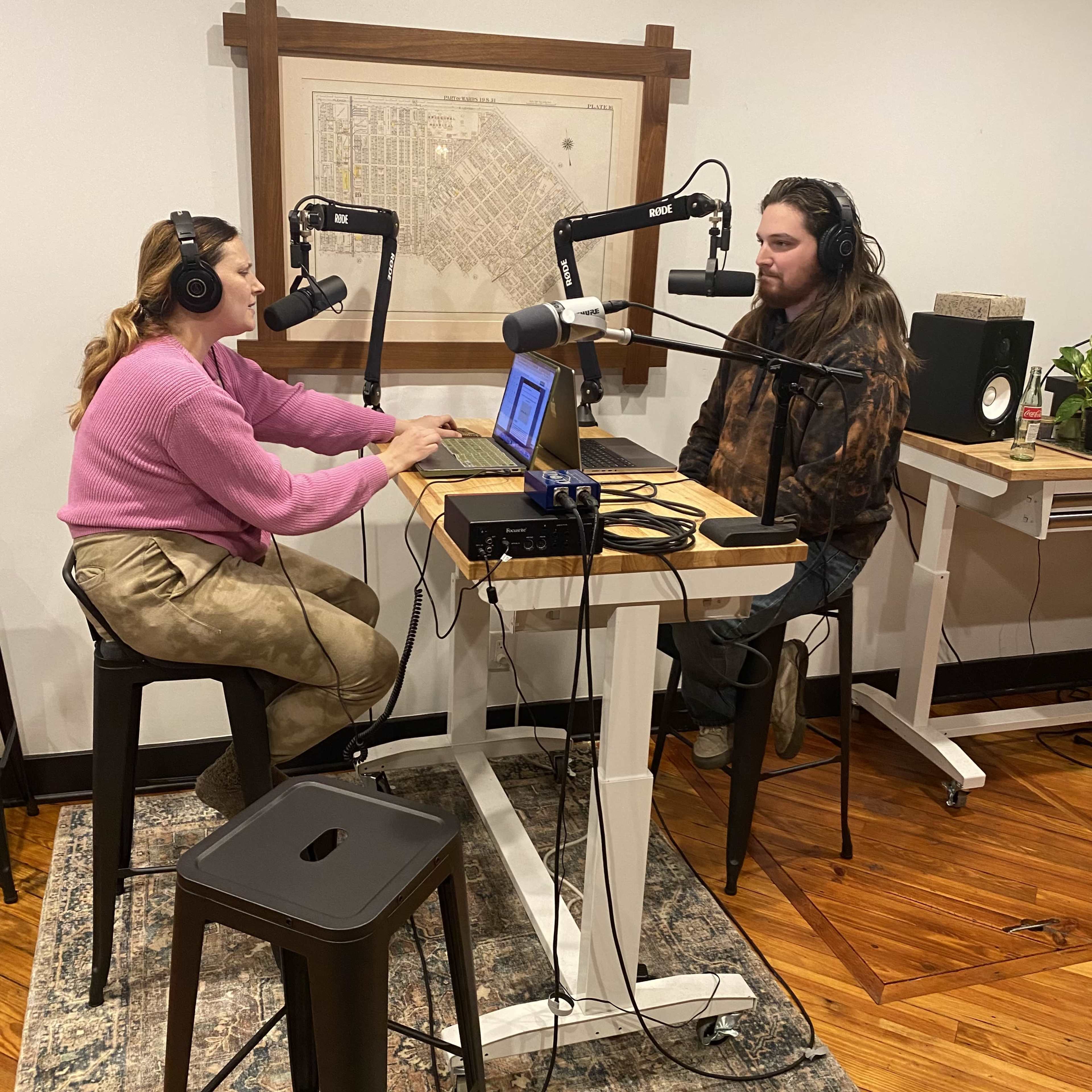 Two people sit at a desk with microphones and laptops, engaging in a podcast recording in a well-lit room.