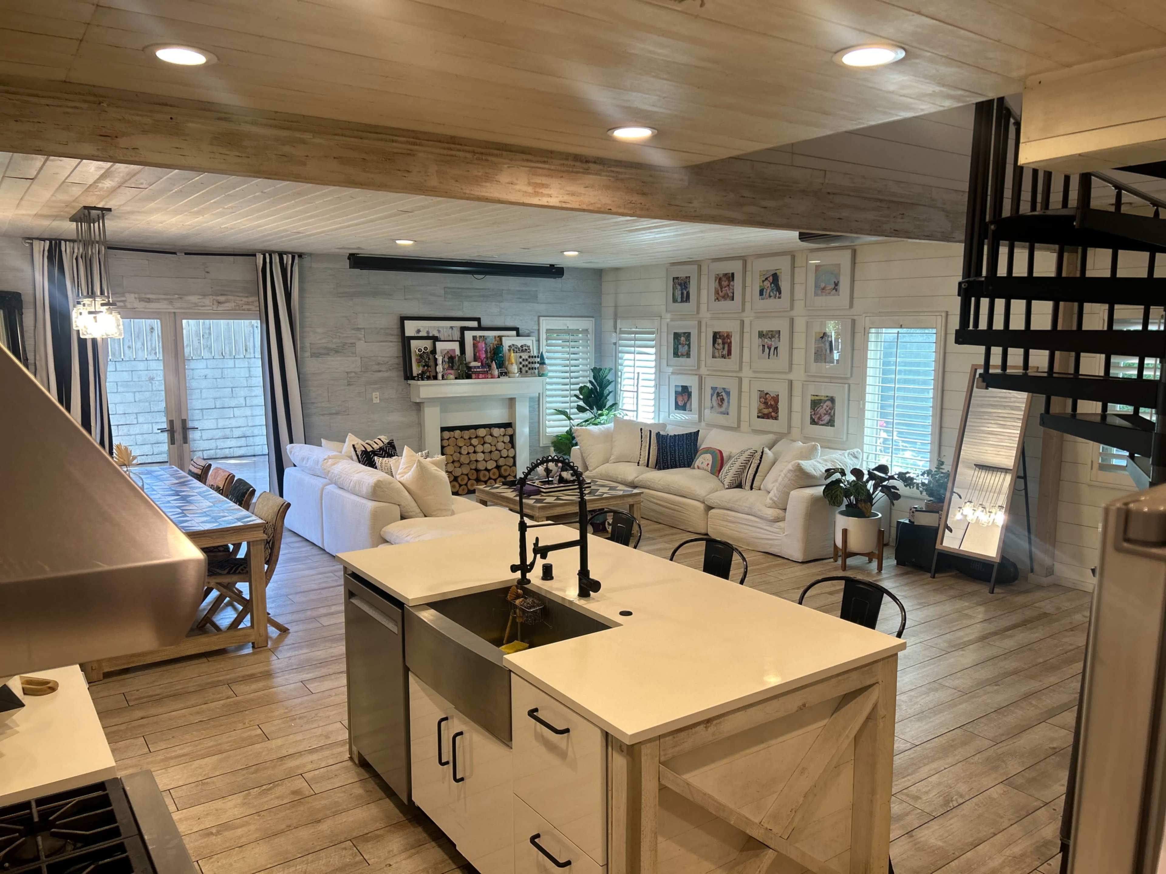 A bright, open-concept living area featuring a living room with a sofa, a dining area with a long table, and a spiral staircase leading to an upper level.