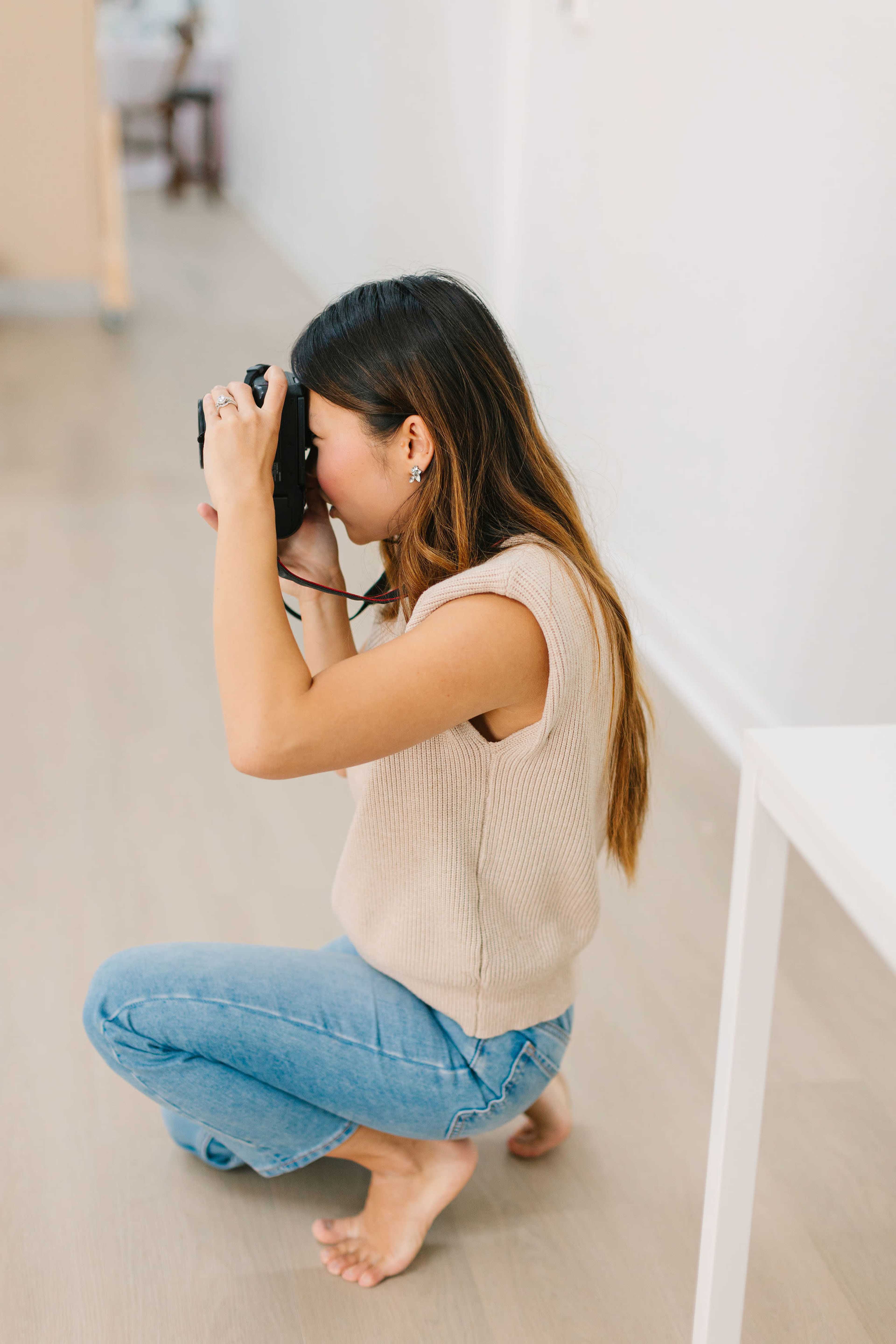 A woman kneels on the floor while taking a photograph with a camera in a brightly lit room.