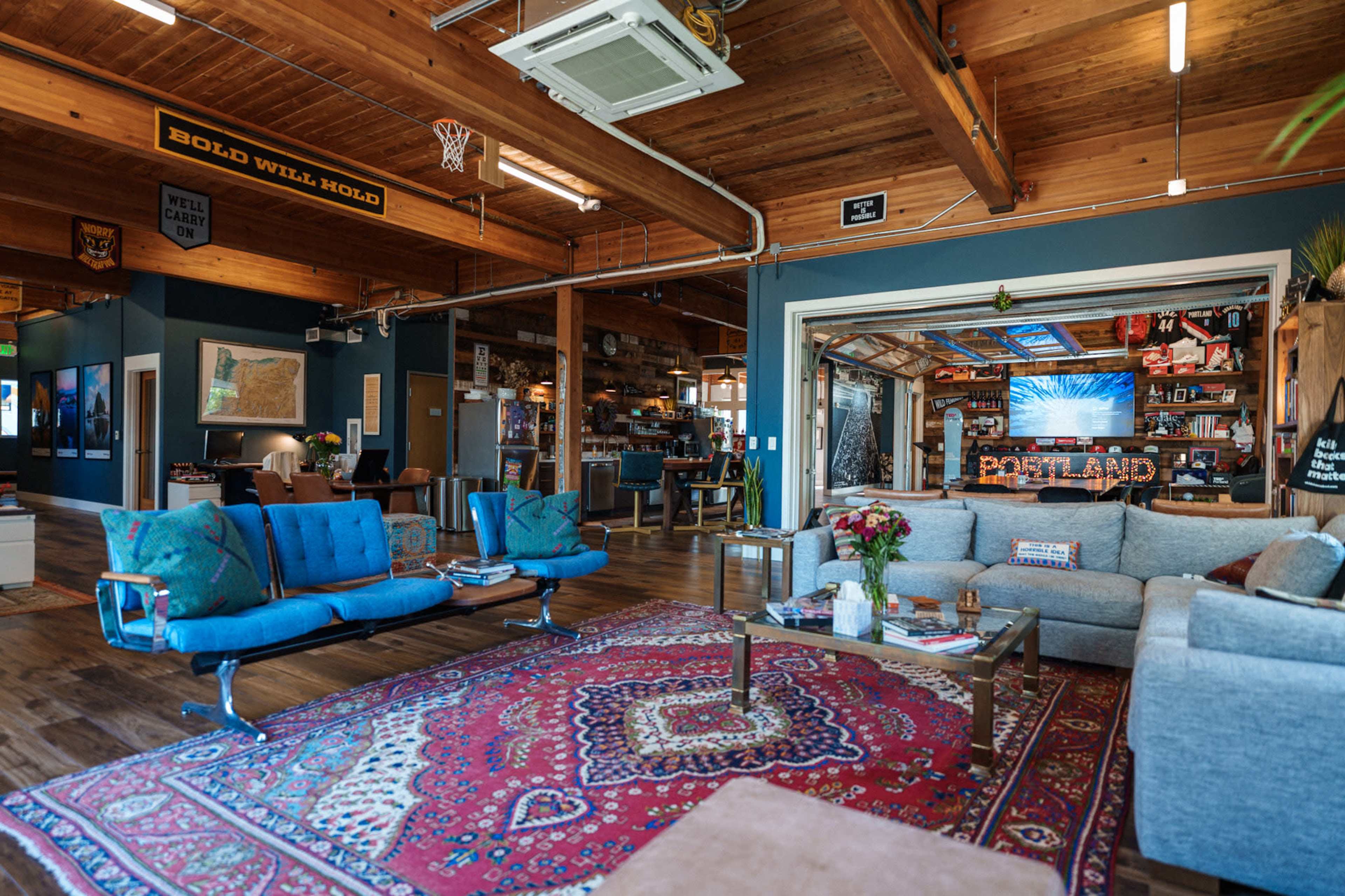 The image shows a spacious, modern living area with wooden ceilings, a large area rug, and various seating options, including a blue couch and a light gray sofa.