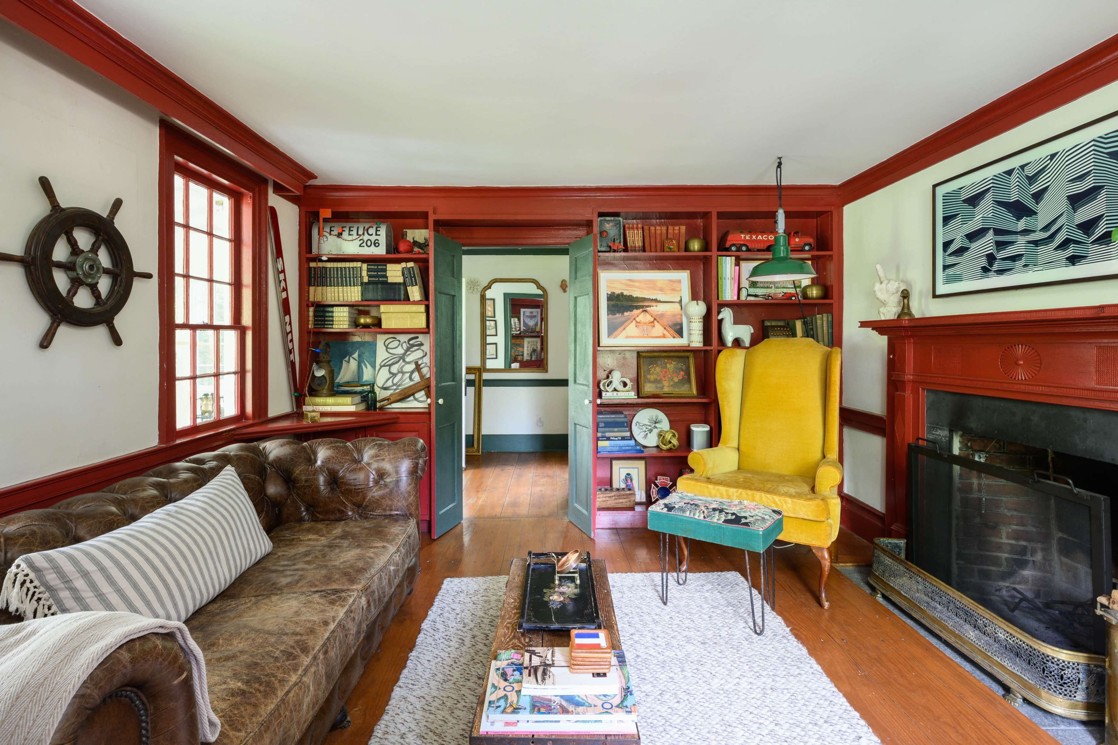 The image shows a cozy living room with a brown leather sofa, a bright yellow armchair, a wooden fireplace, and shelves filled with books and decorative items.