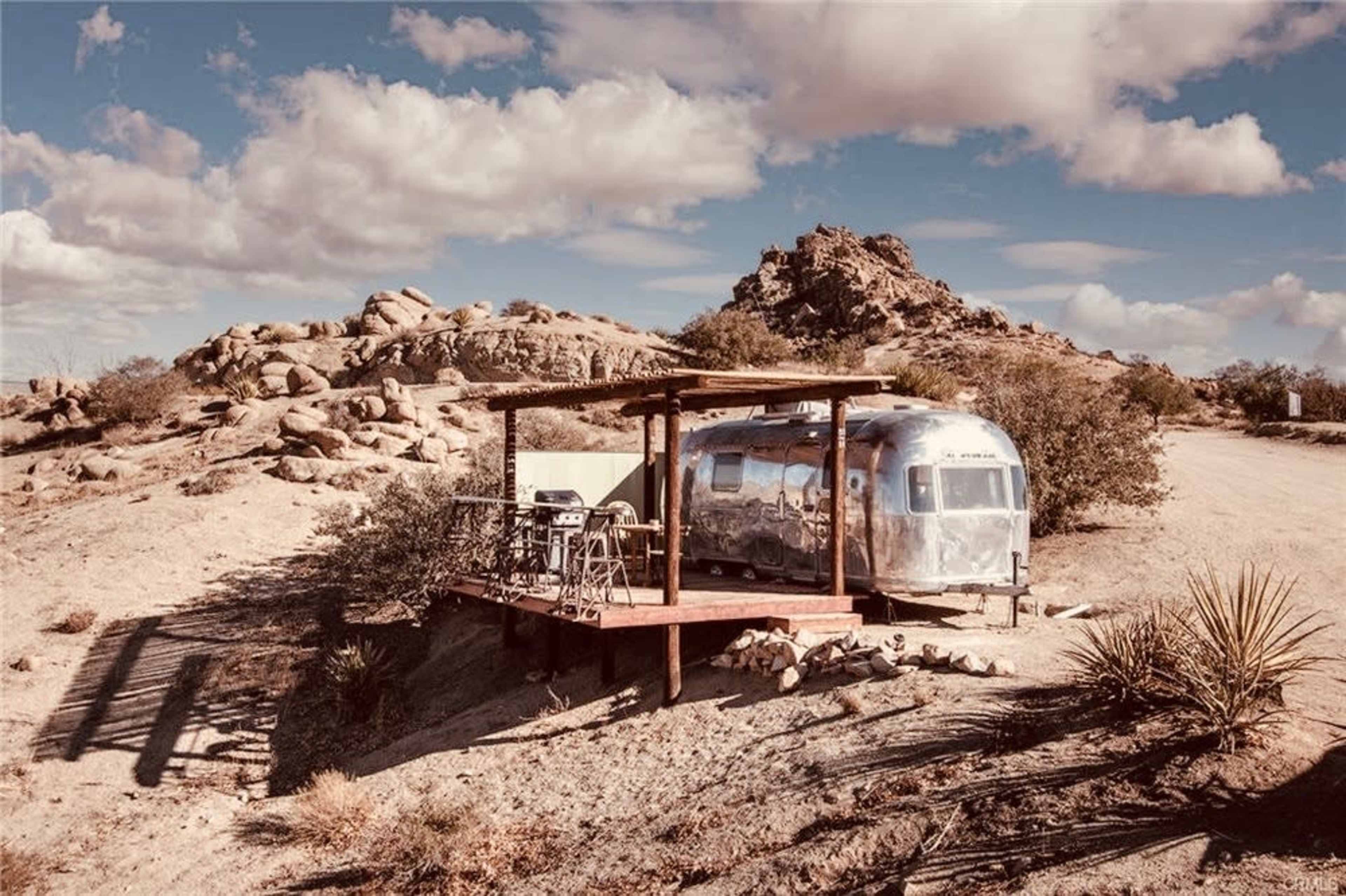 An Airstream trailer is stationed on a wooden deck amidst rocky terrain and sparse vegetation under a cloudy sky.
