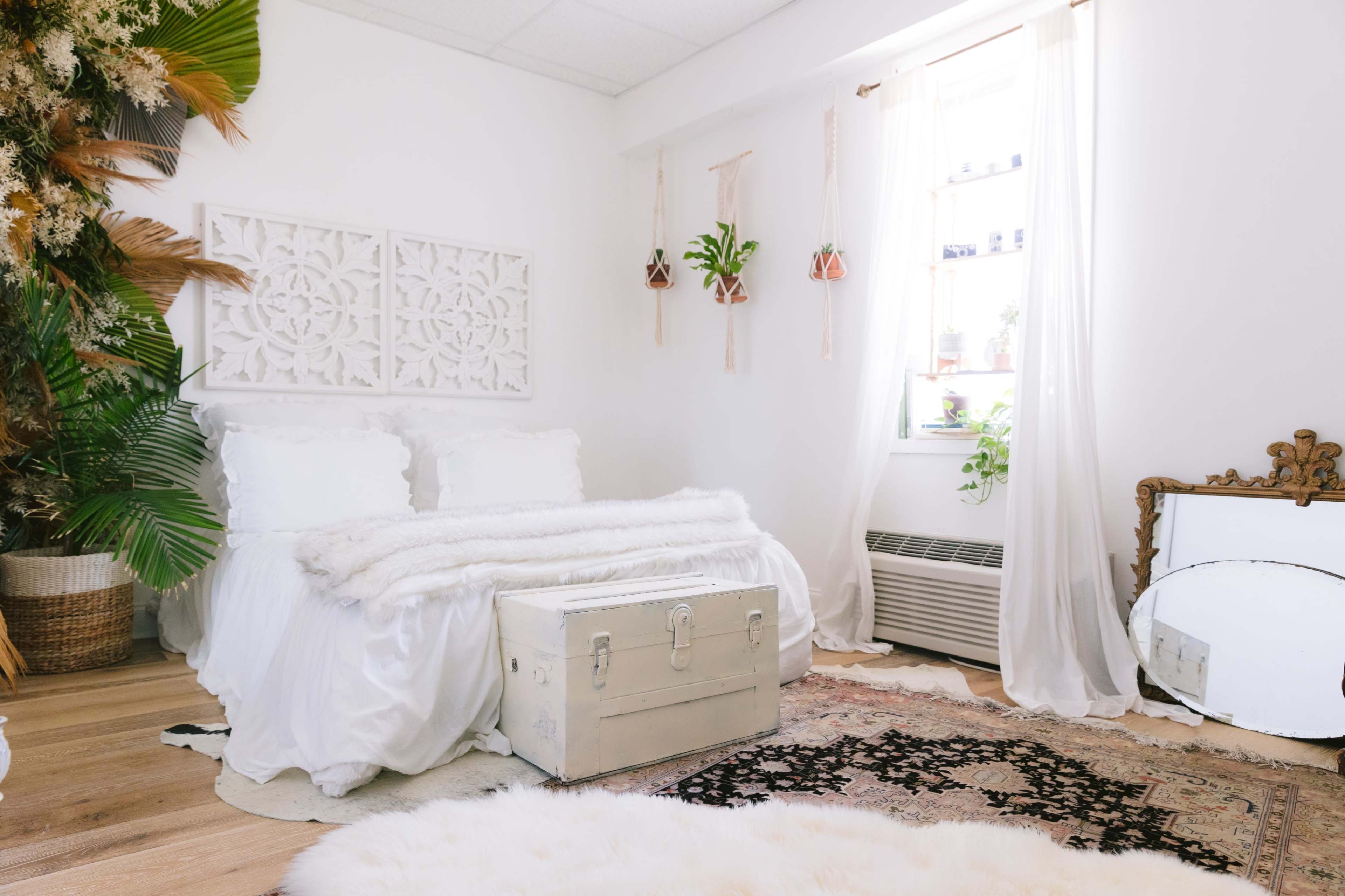 The image shows a bright, minimalist bedroom featuring a white bed with fluffy bedding, a vintage trunk at its foot, and plants positioned by a large window, with a decorative mirror on the floor.