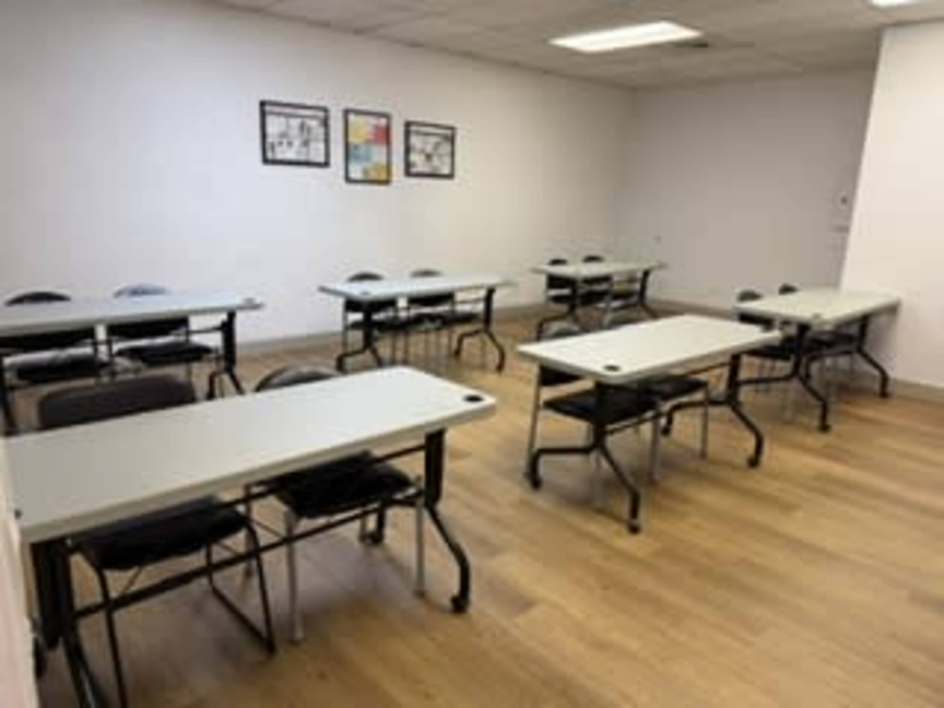 A clean, empty classroom features several tables and chairs arranged neatly with minimal decorations on the walls.