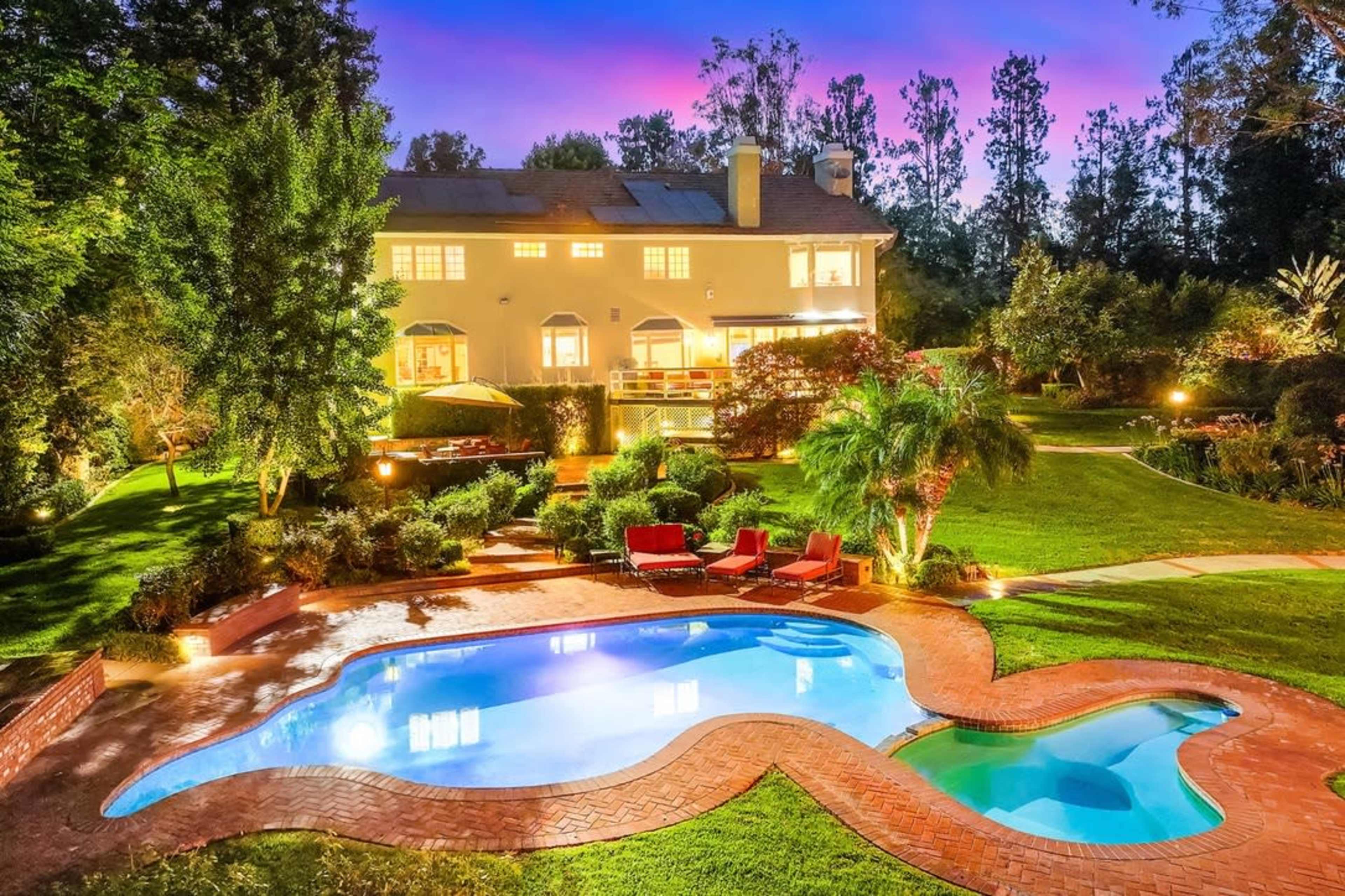 The scene features a well-lit backyard with a swimming pool shaped like a figure, surrounded by lush greenery, lounge chairs, and a two-story house in the background.