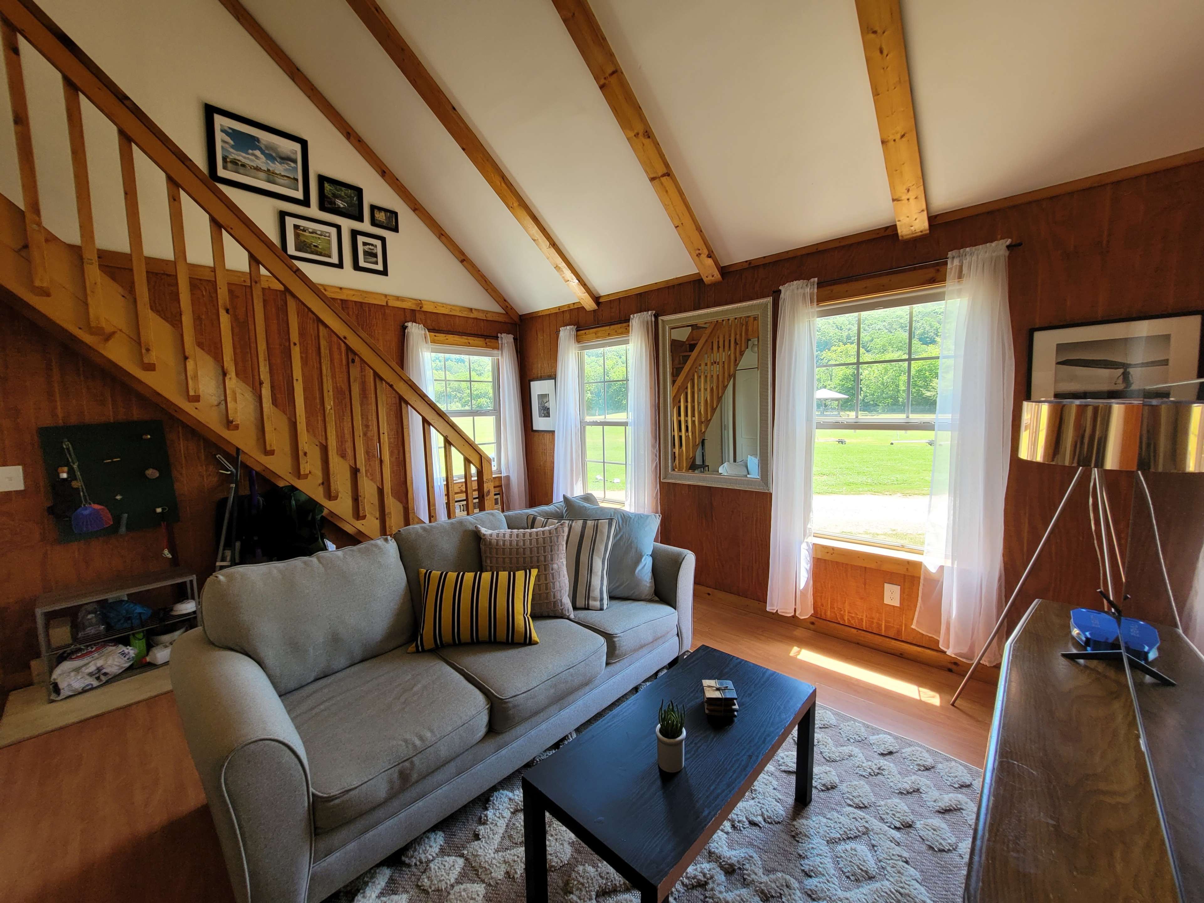The interior of a cozy living room features a gray sofa with decorative pillows, a coffee table, and large windows that offer views of a green outdoor landscape.