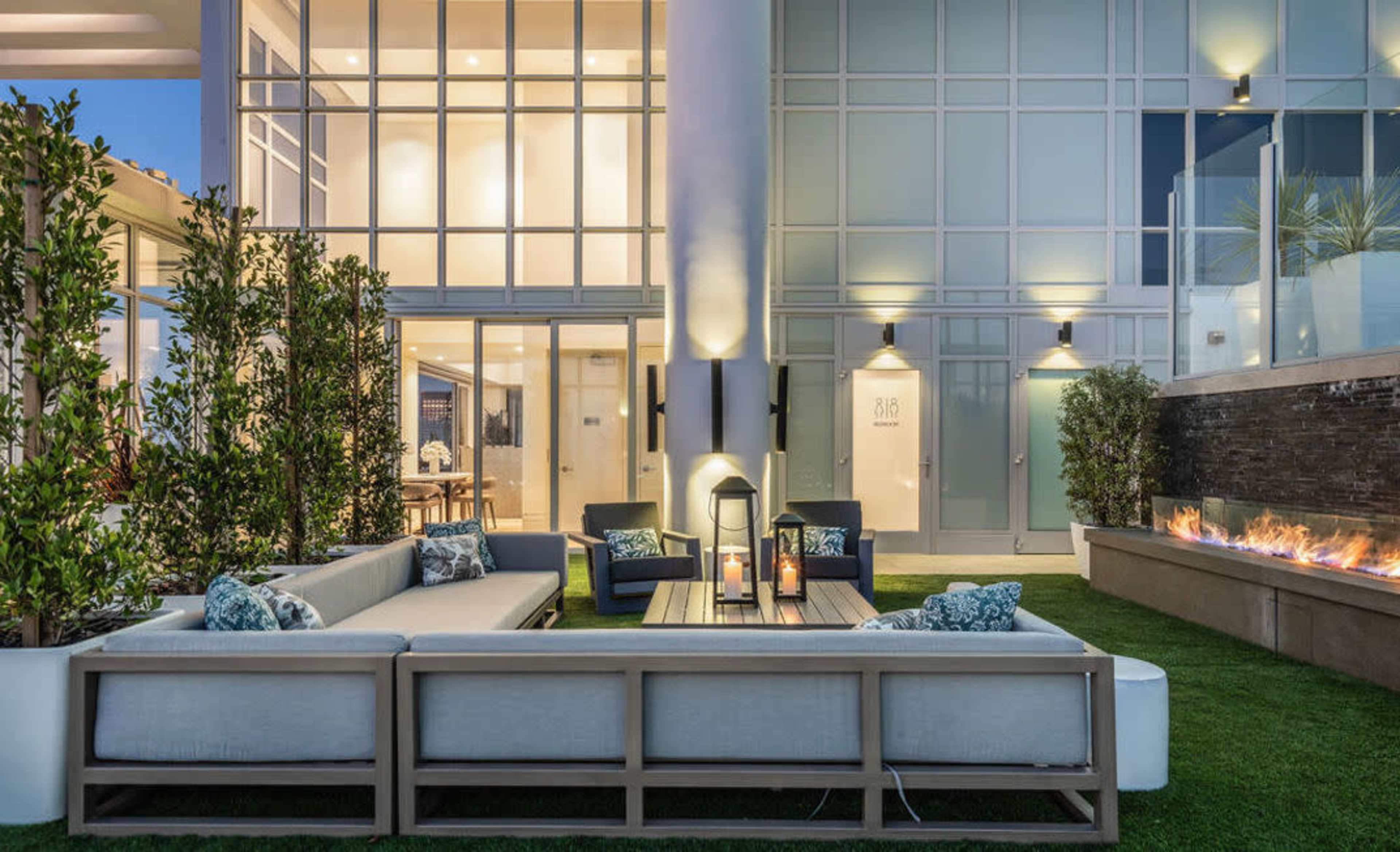 The image shows a modern outdoor seating area with a sofa, coffee table, and lanterns, surrounded by greenery and a gas fire feature.