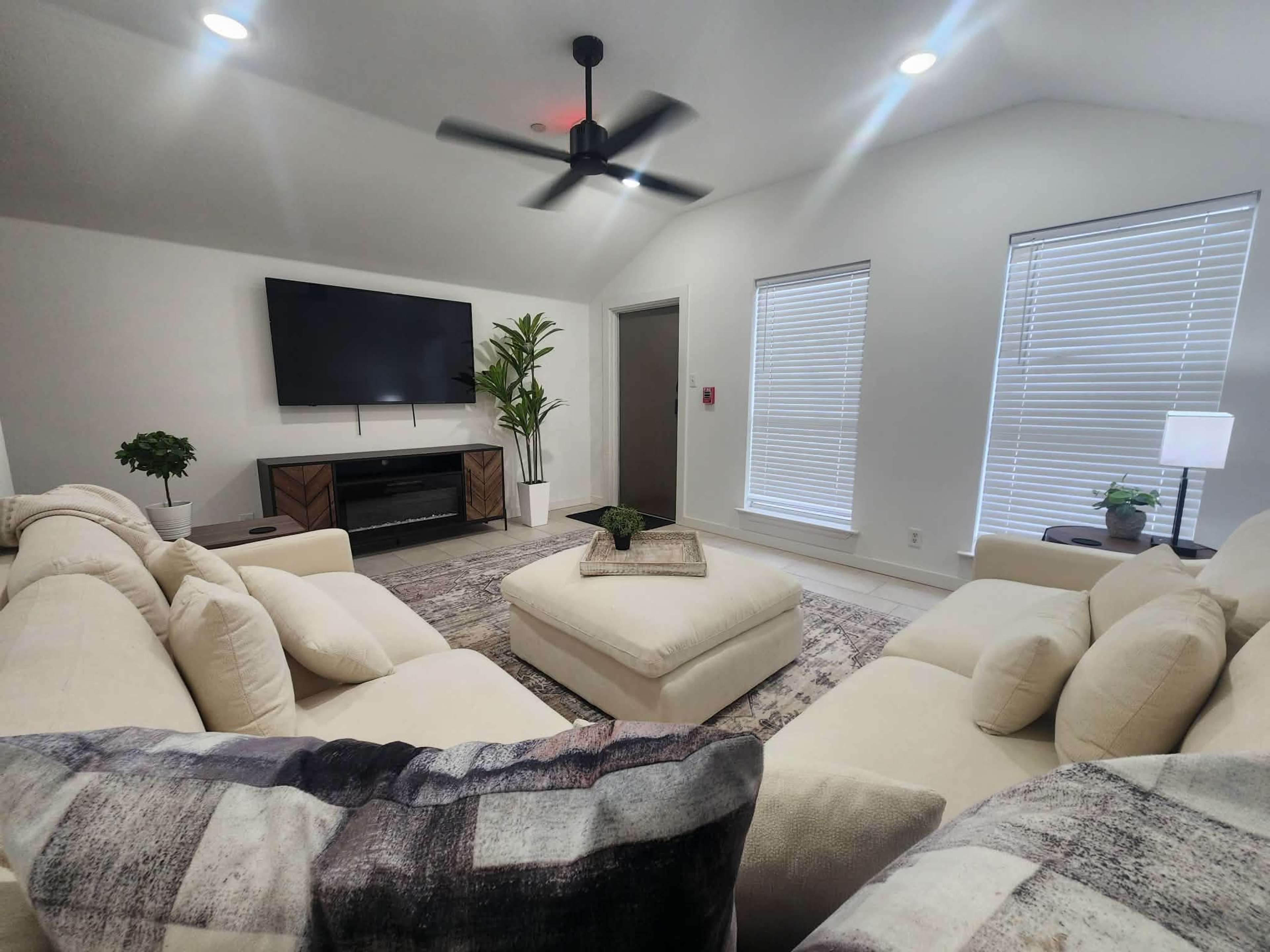 The image shows a modern living room with a light-colored sectional sofa, a large ottoman in the center, a wall-mounted television, and natural light coming through two window blinds.