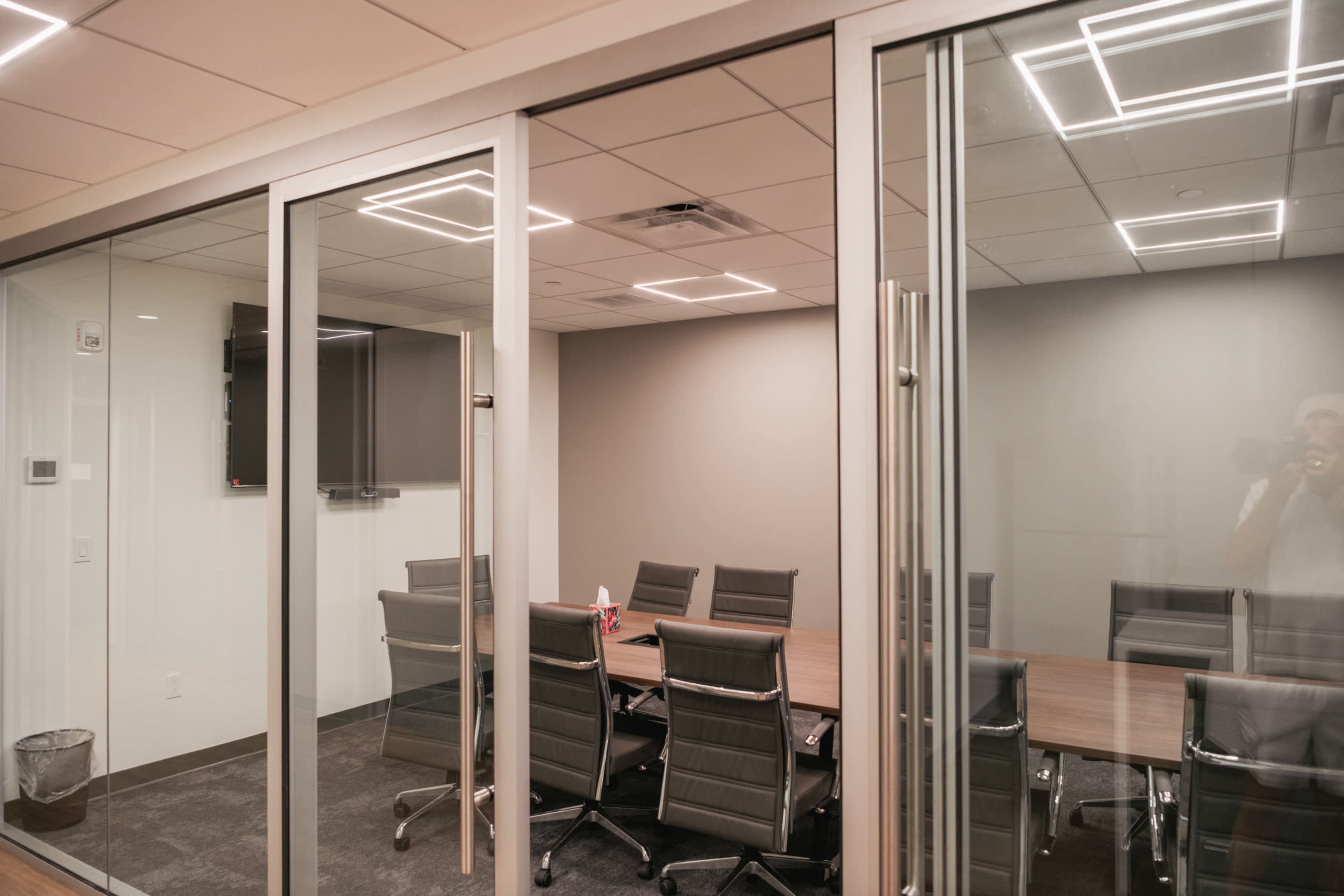 The image shows a modern conference room with a large table surrounded by chairs, enclosed by glass walls and a door.