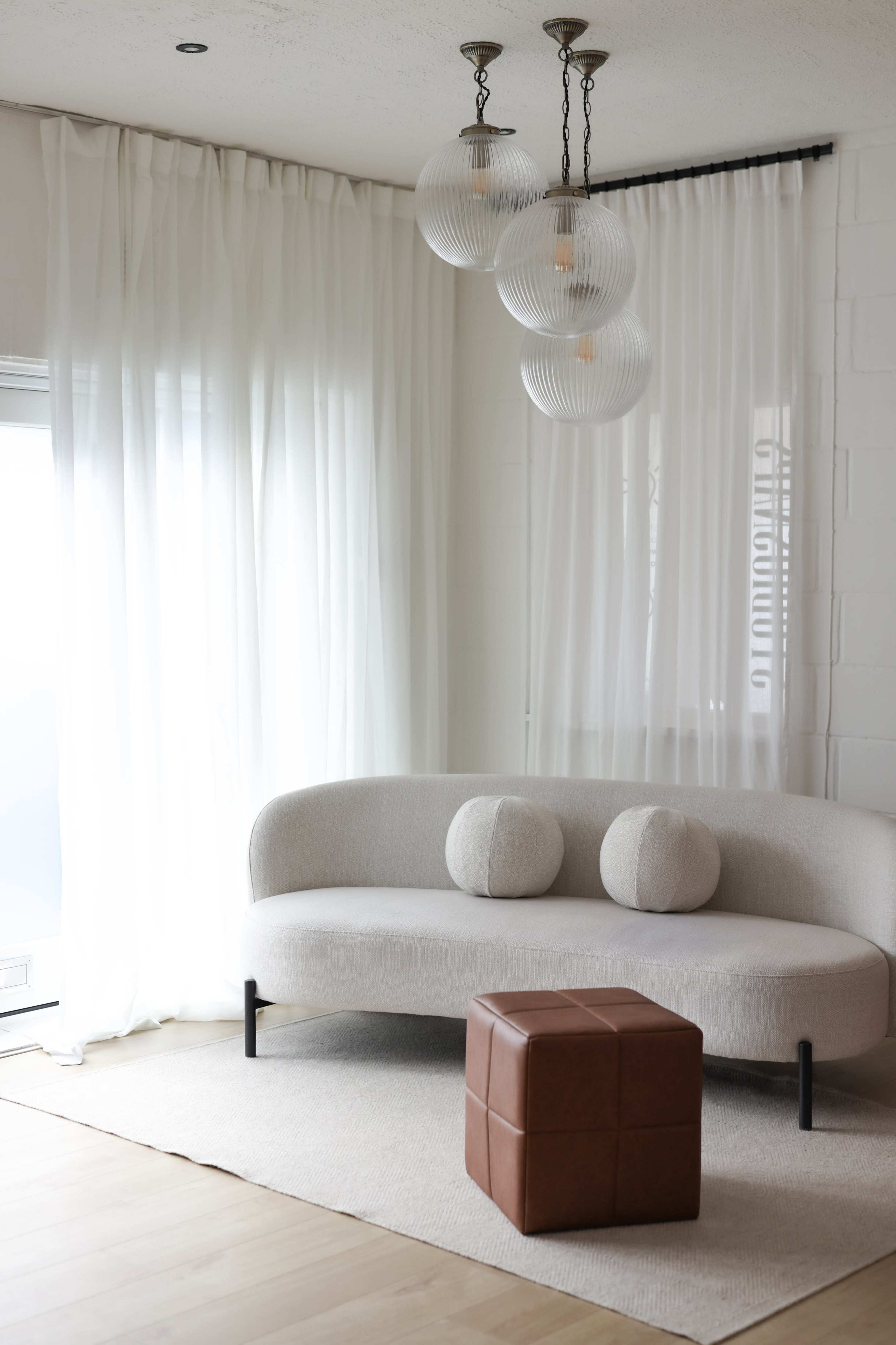 The image shows a modern living area featuring a curved beige sofa with two round cushions and a brown leather ottoman, illuminated by three pendant lights above.
