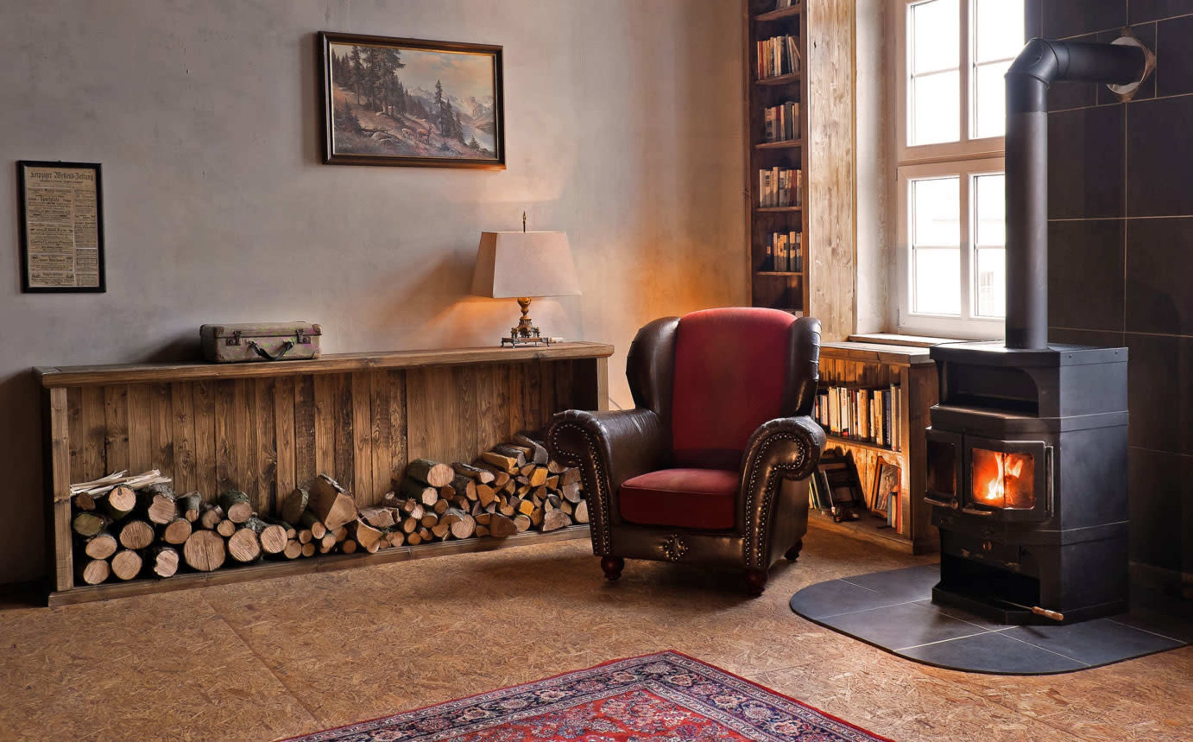 The image depicts a cozy room featuring a wood stove, a red armchair, a stack of firewood, and bookshelves filled with books.