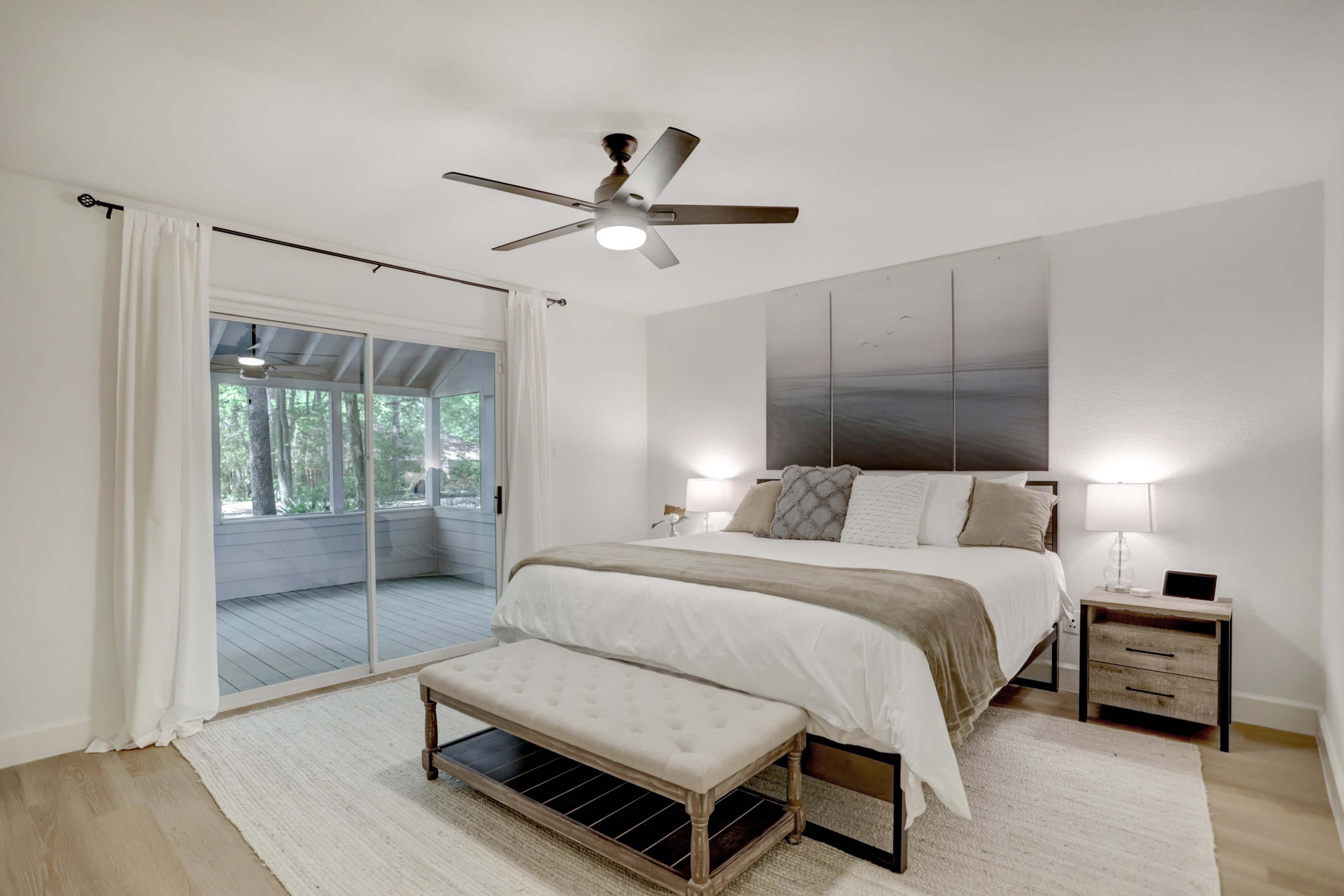 A modern bedroom features a bed with a light-colored duvet, a bench at the foot, and sliding glass doors leading to a deck surrounded by trees.