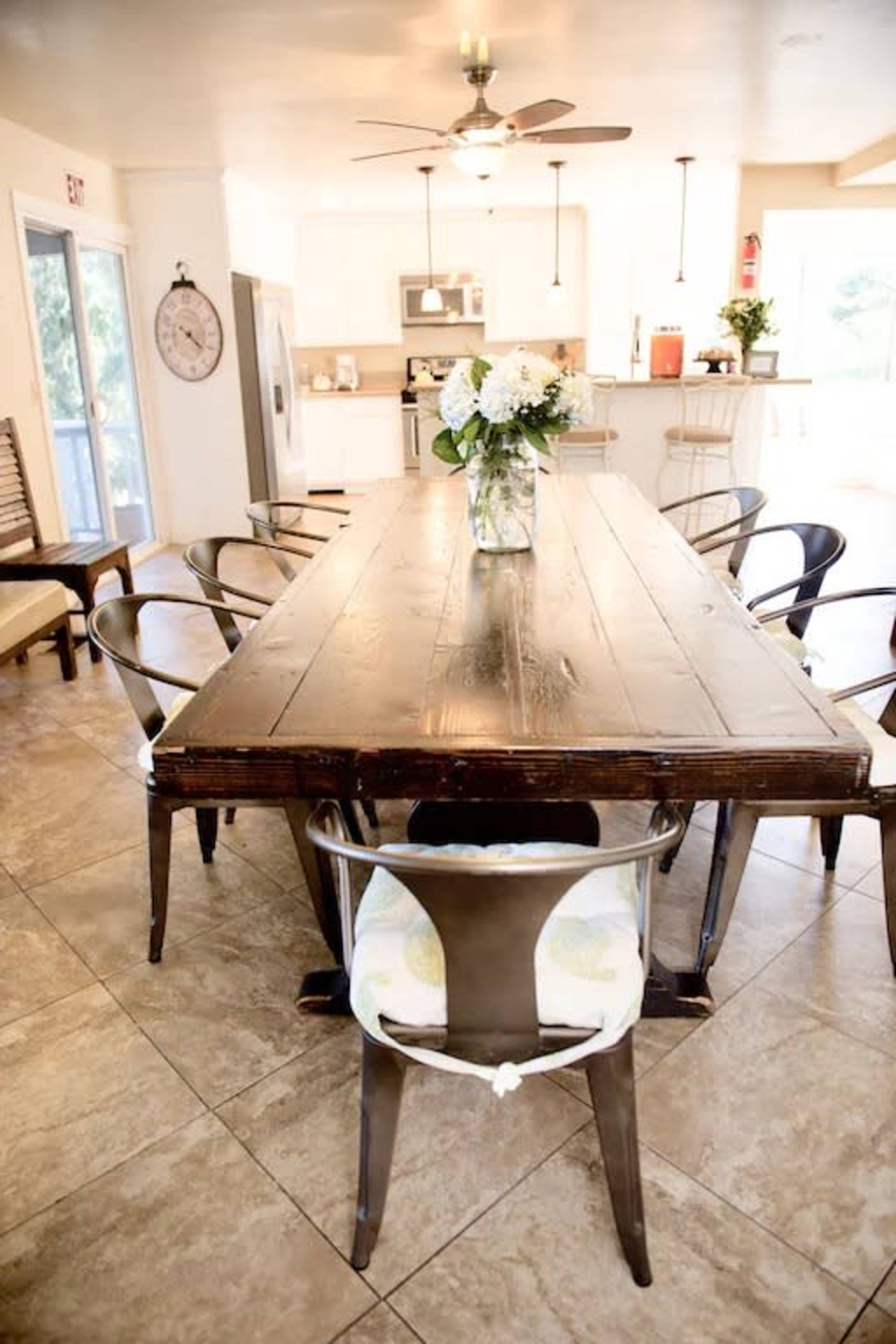 The image shows a spacious dining area with a large wooden table surrounded by metal and wooden chairs, and a vase of flowers as the centerpiece.