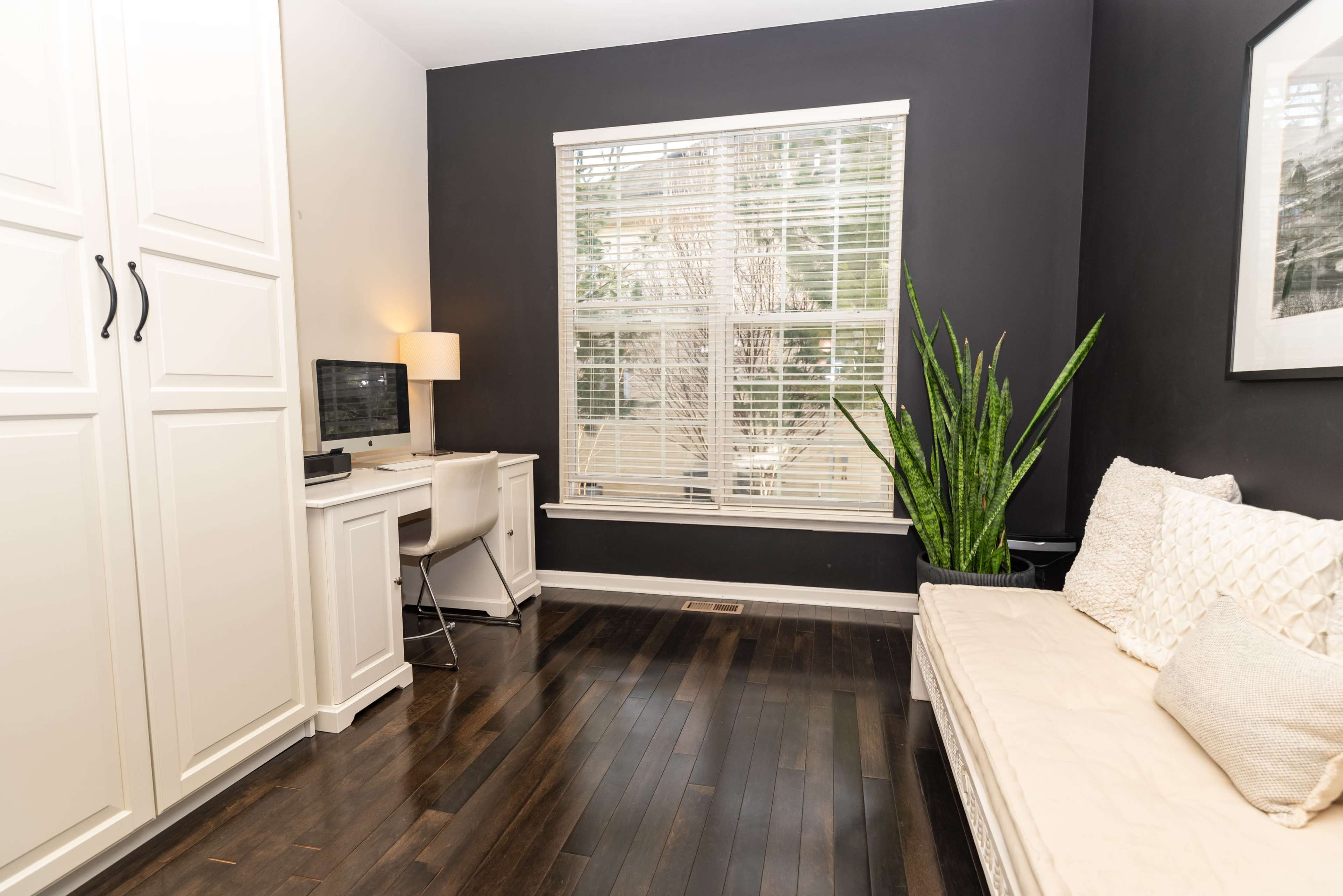 A small home office features a desk with a computer, a white chair, a plant in the corner, and a couch along one wall, all within a room accented by dark walls and a large window.