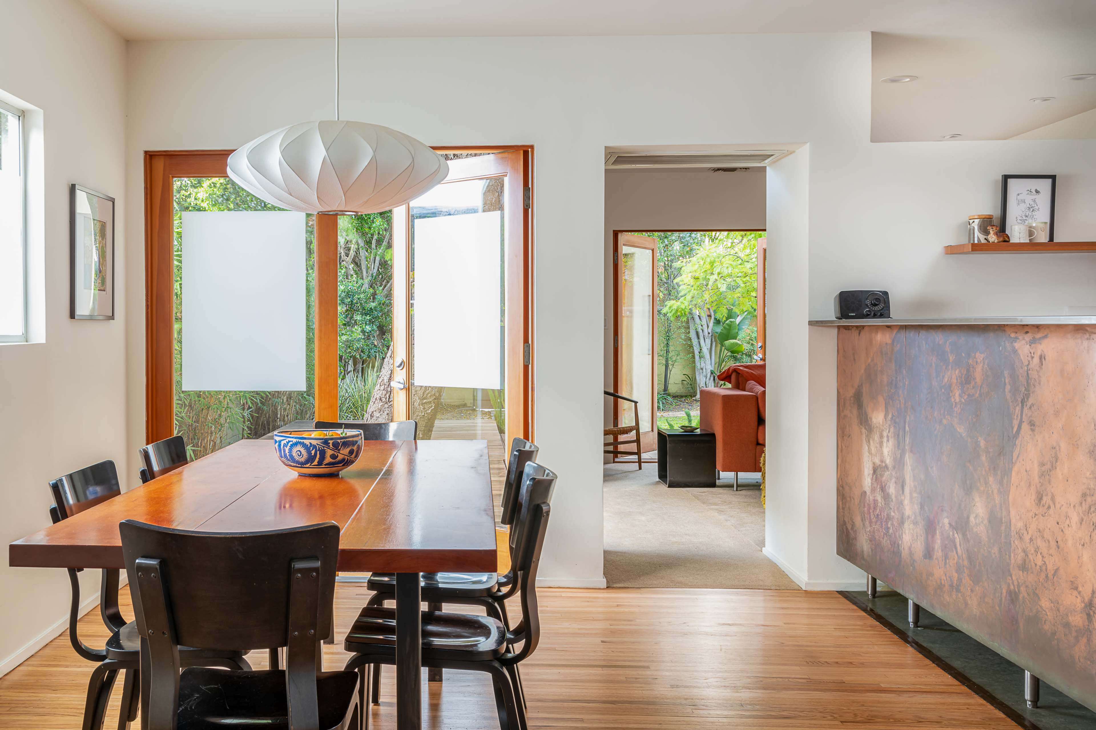 A dining area with a wooden table and black chairs opens into a sunny, green outdoor space through sliding glass doors.
