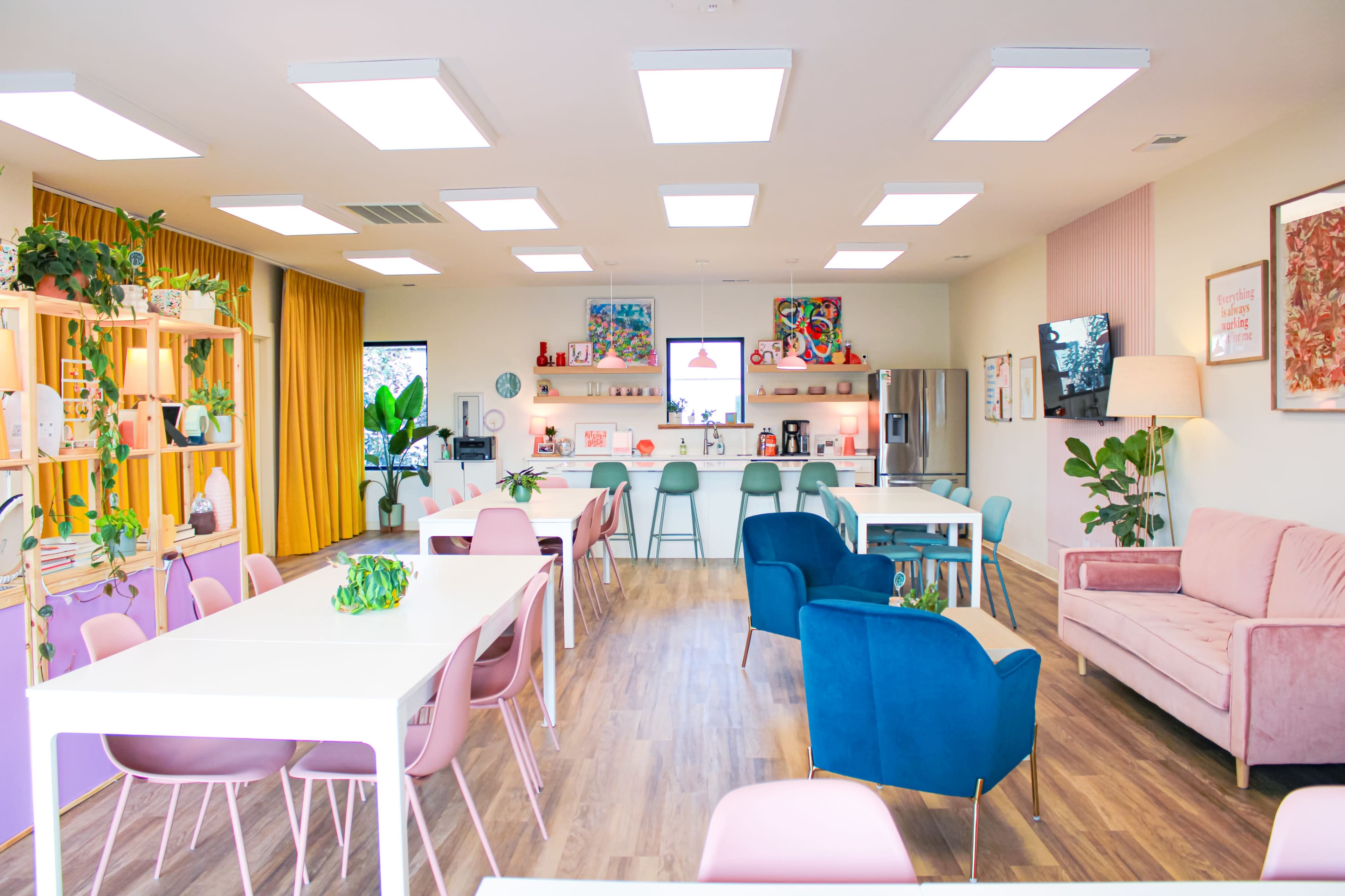 A brightly lit modern interior of a café or communal space with pink and blue furniture, including tables and chairs, as well as shelves displaying decorative items.