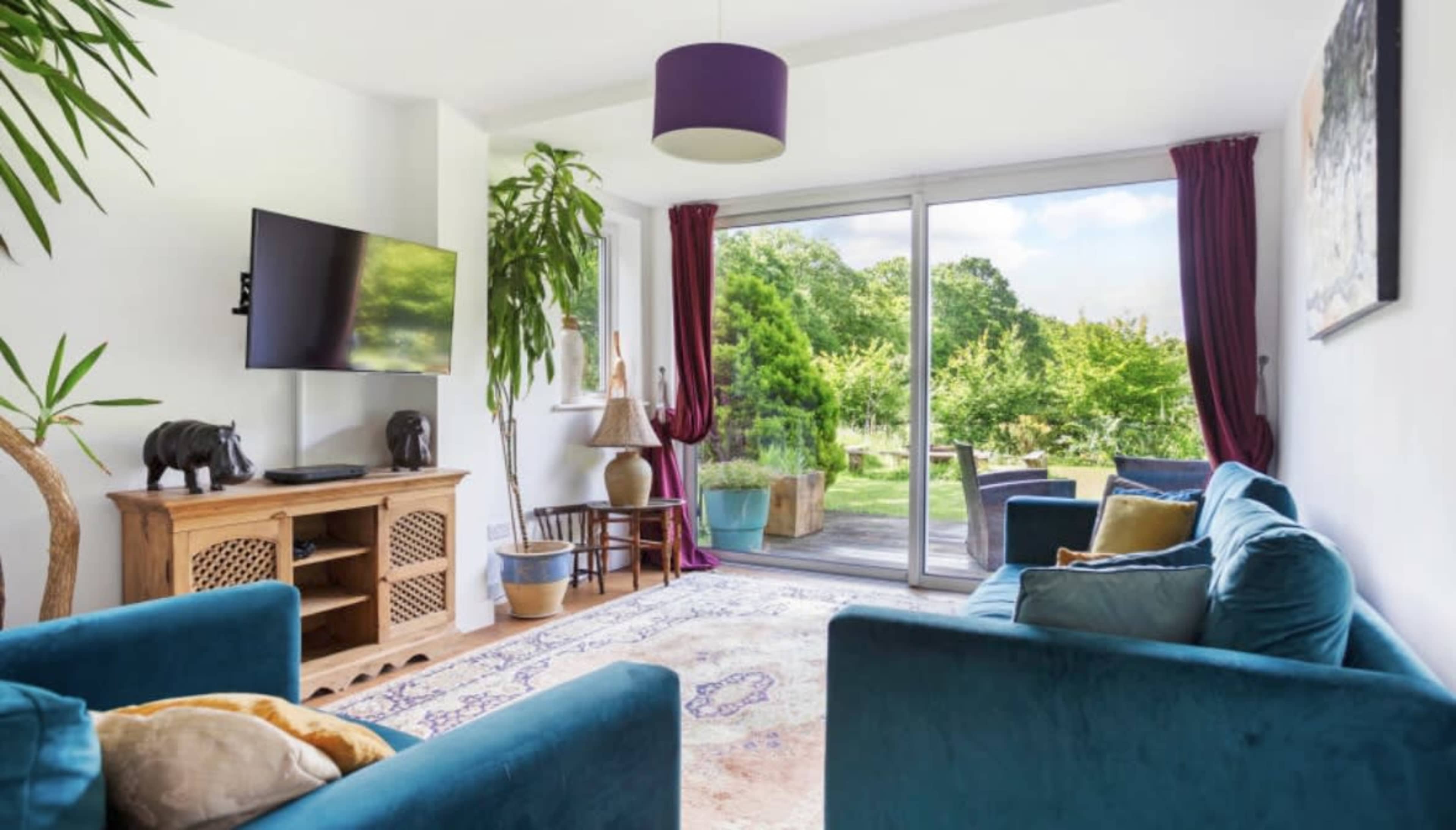 A cozy living room with teal sofas, a television mounted on the wall, and glass doors leading to a lush garden.
