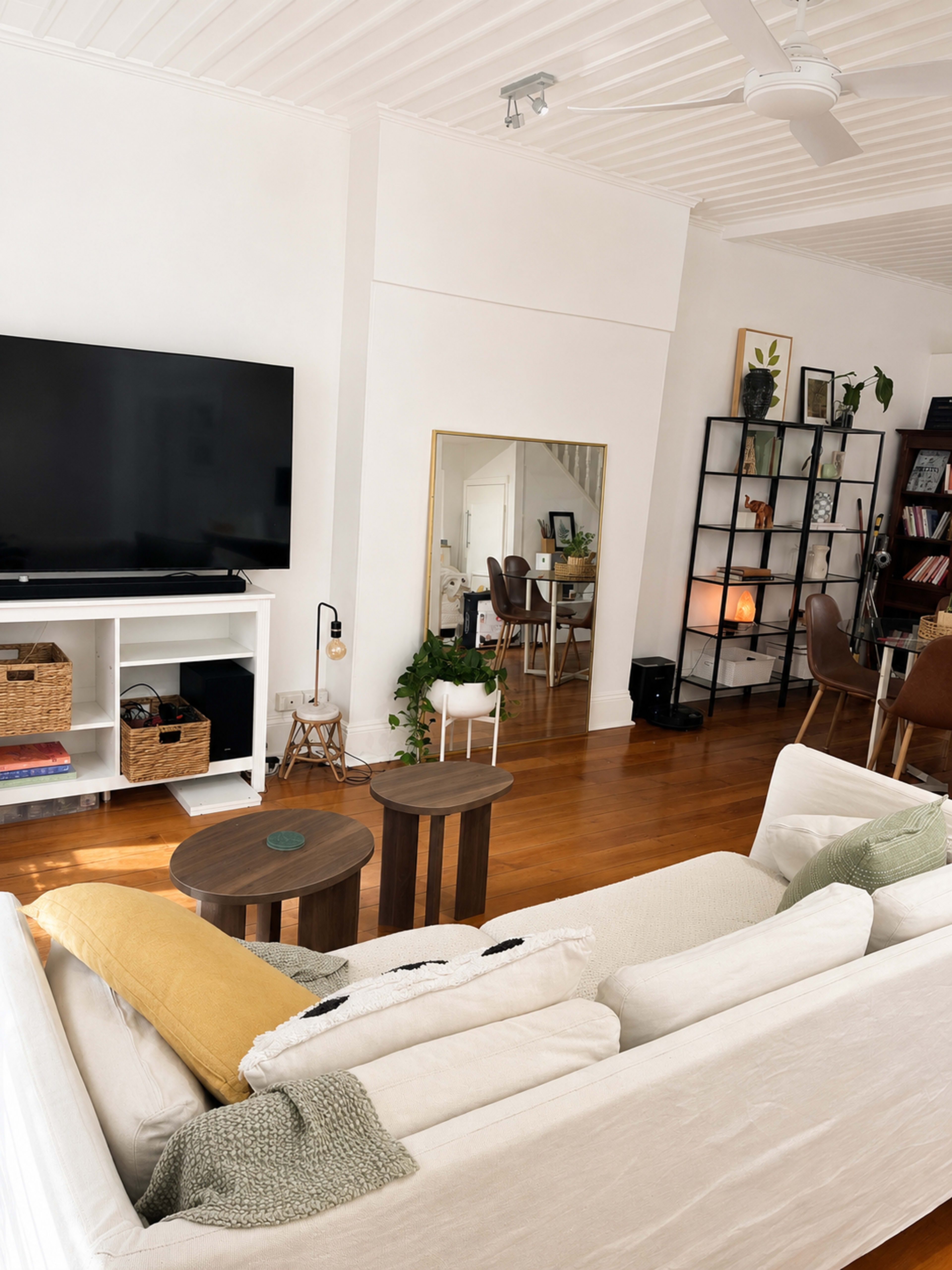 The image depicts a modern living room with a white couch, a large television on a media console, and a mirror reflecting a bookshelf and dining area.