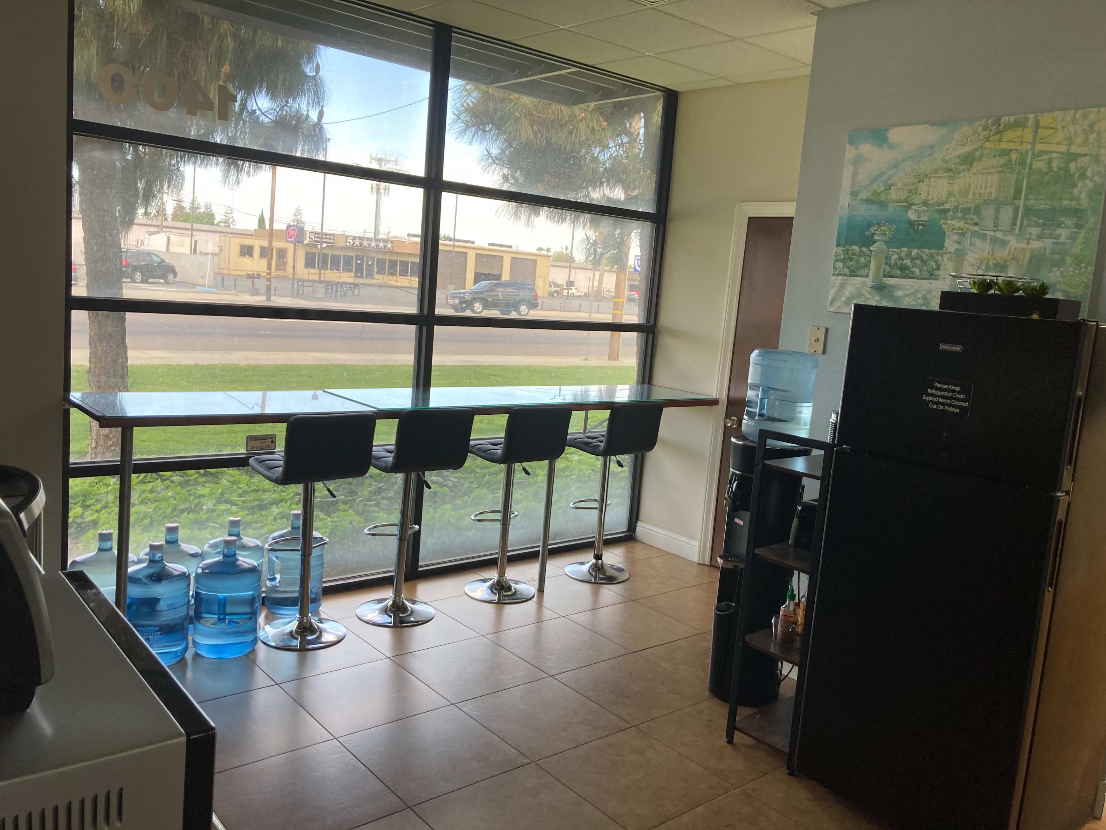 A corner of a room with a large window overlooking a street, featuring a bar-style table with four high stools and a refrigerator beside water dispensers.