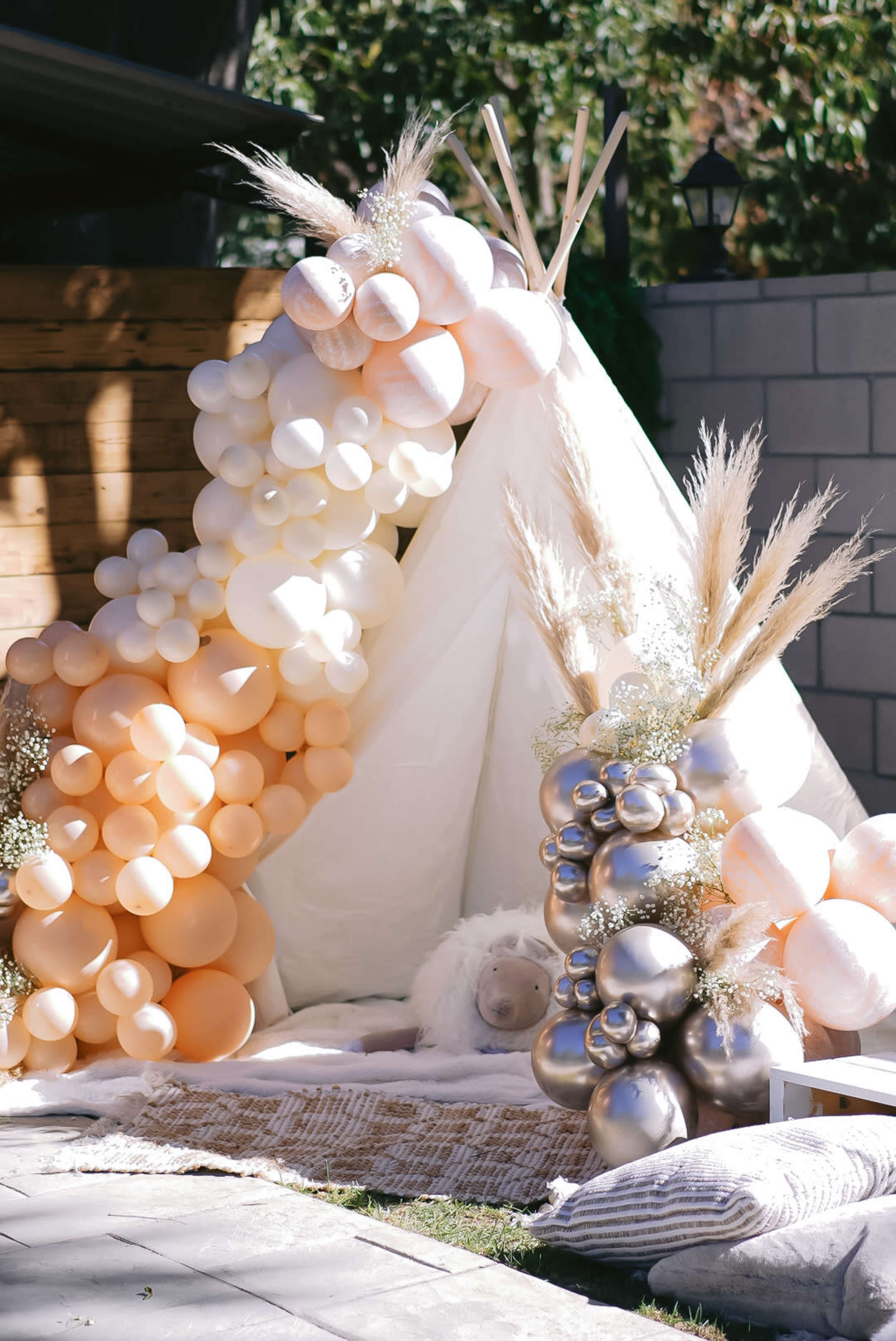 A decorative area with a teepee made of white fabric, surrounded by clusters of colored balloons and pampas grass.
