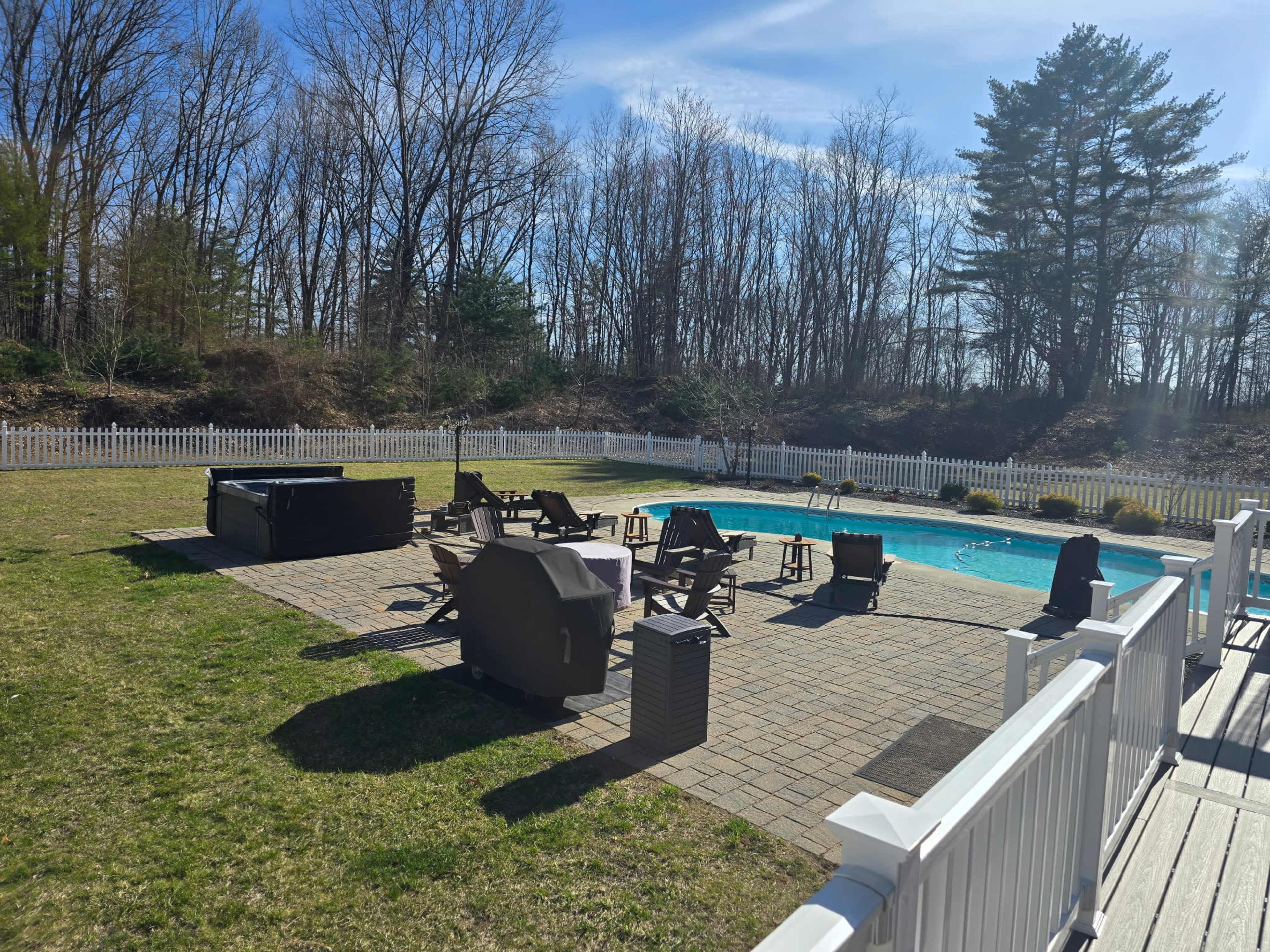 The image shows a backyard pool area with a paved patio, several lounge chairs, a hot tub, and a white picket fence, surrounded by bare trees under a clear blue sky.