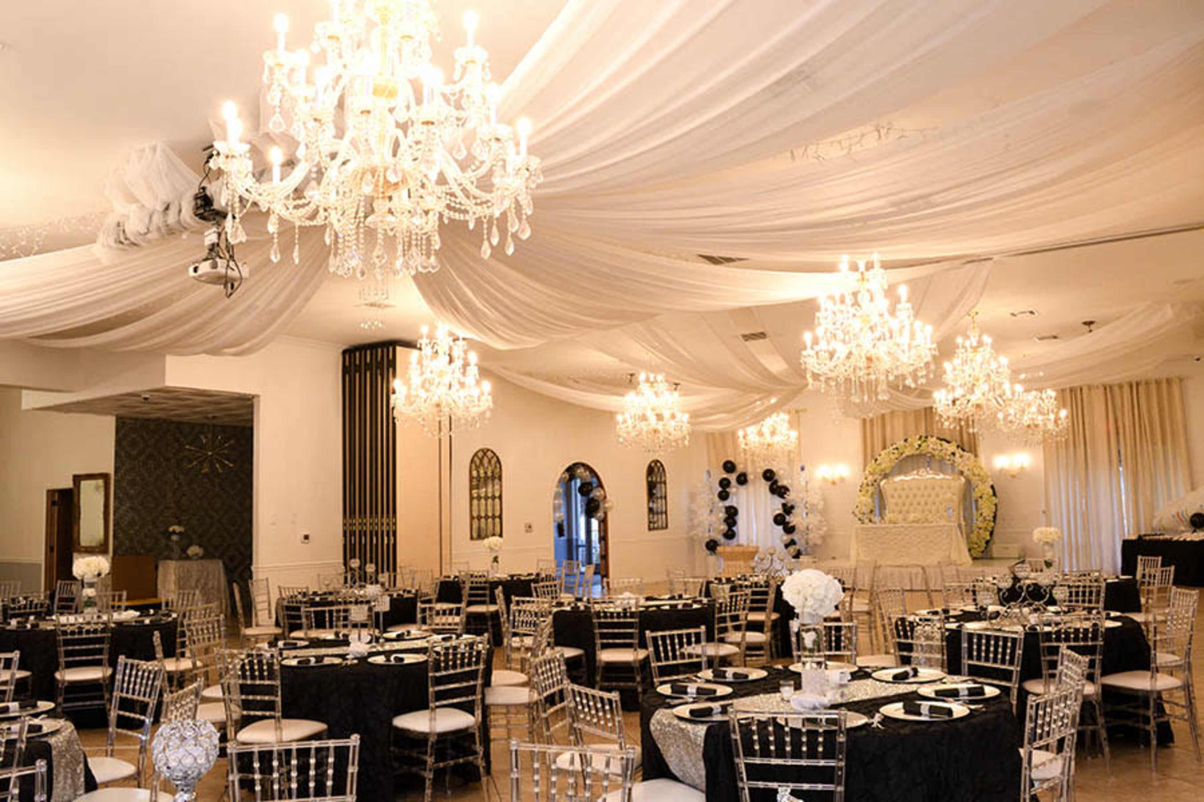An elegantly decorated event space with multiple chandeliers, draped ceiling fabric, and round tables set for a formal gathering.