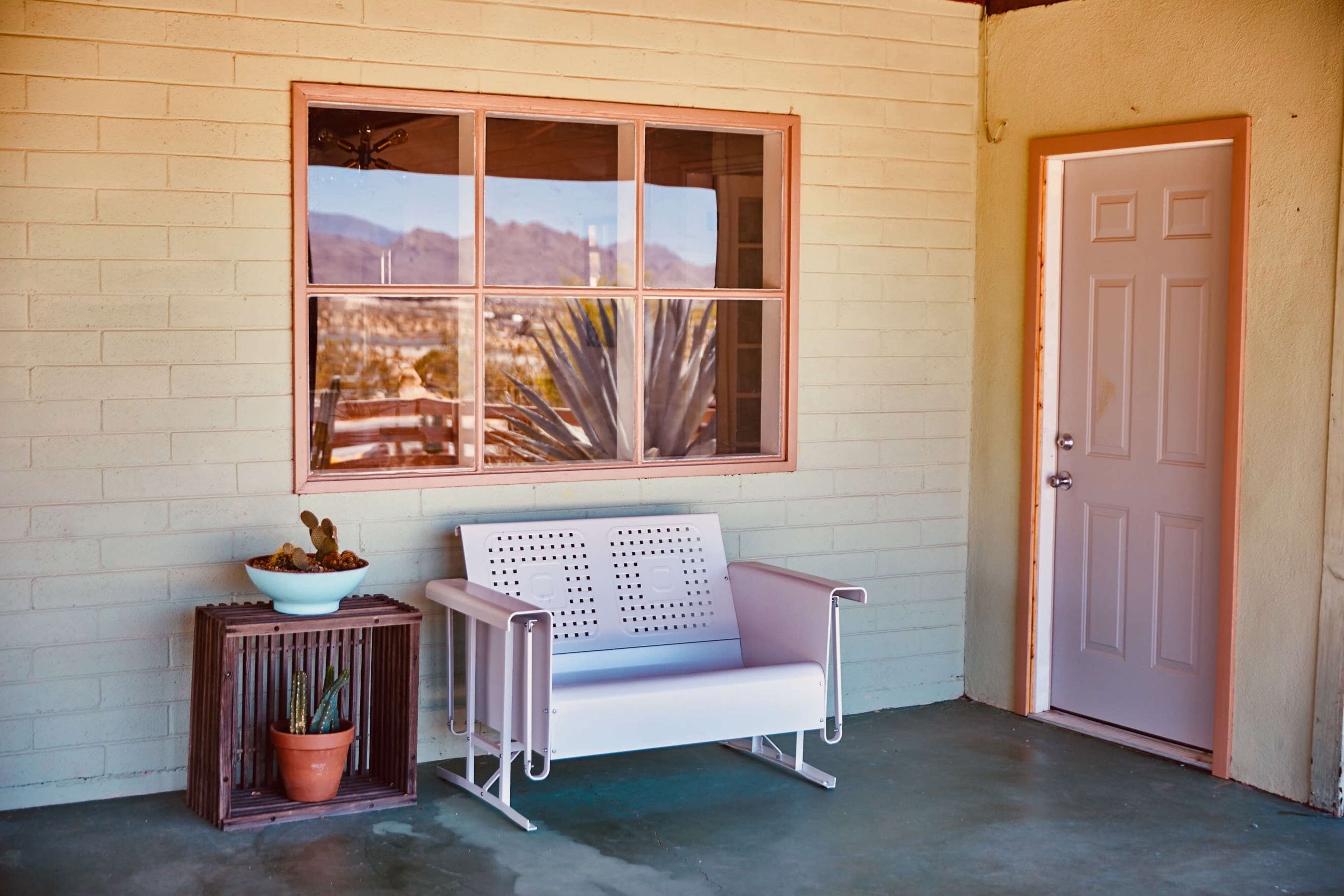 The image shows a porch area featuring a white bench, a small plant pot, and a door with a window reflecting mountains in the background.