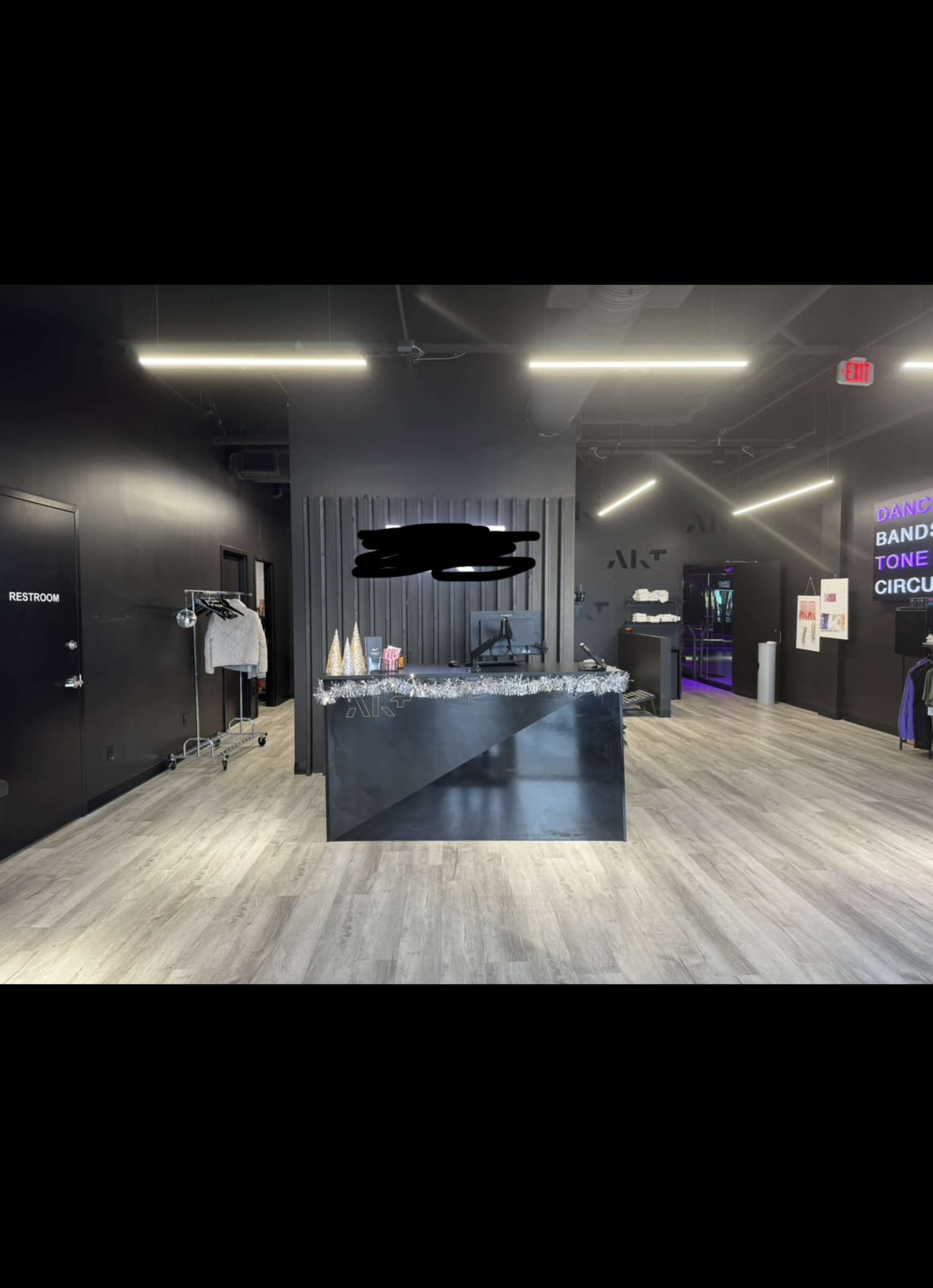 A modern reception area with a sleek black front desk, grey wood-like flooring, and various items displayed on the counter, along with a clothing rack and restroom sign visible.