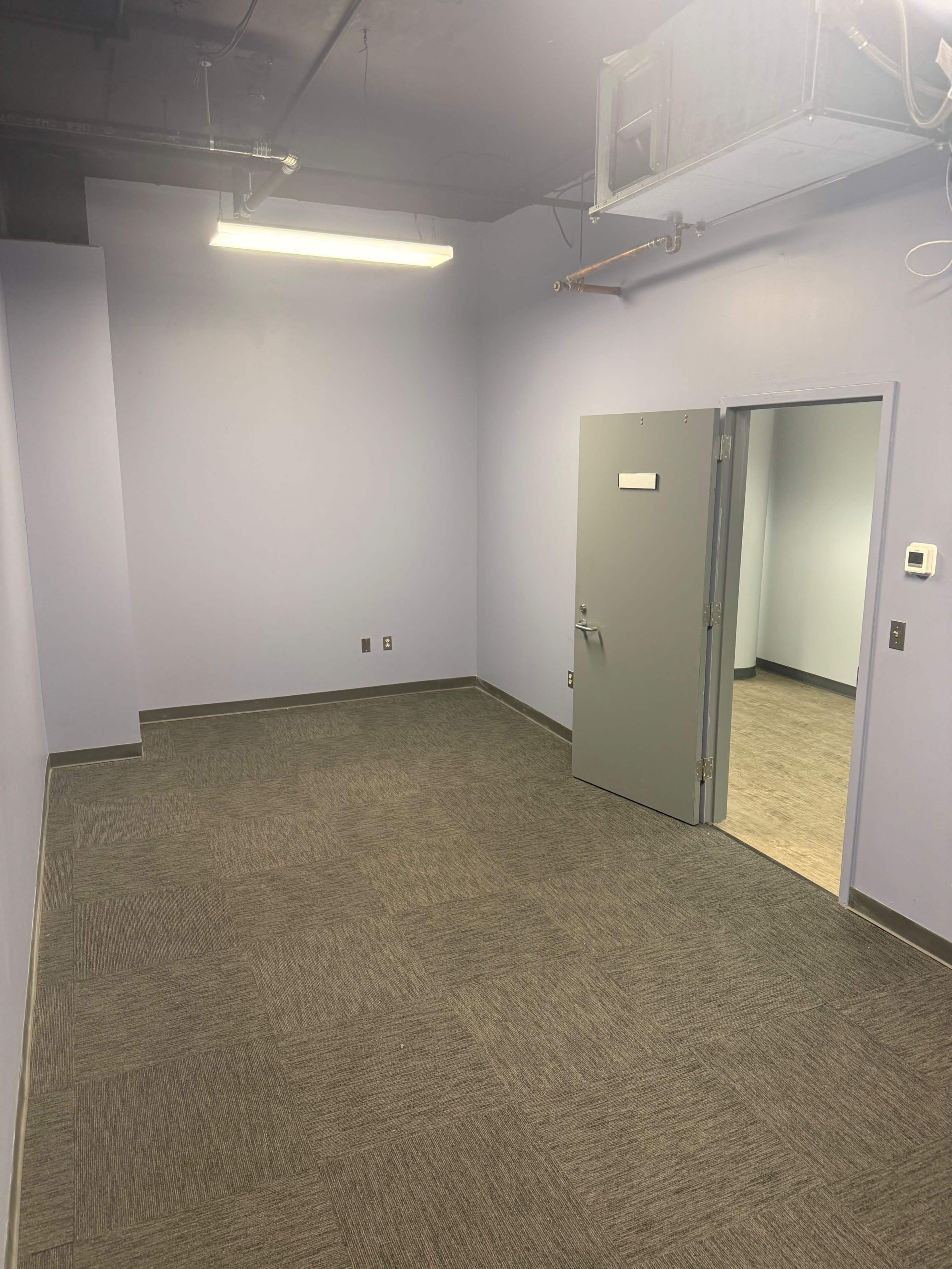The image shows an empty, sparsely furnished room with light purple walls and gray carpeting, featuring a door on the left and another door leading to an adjacent space.