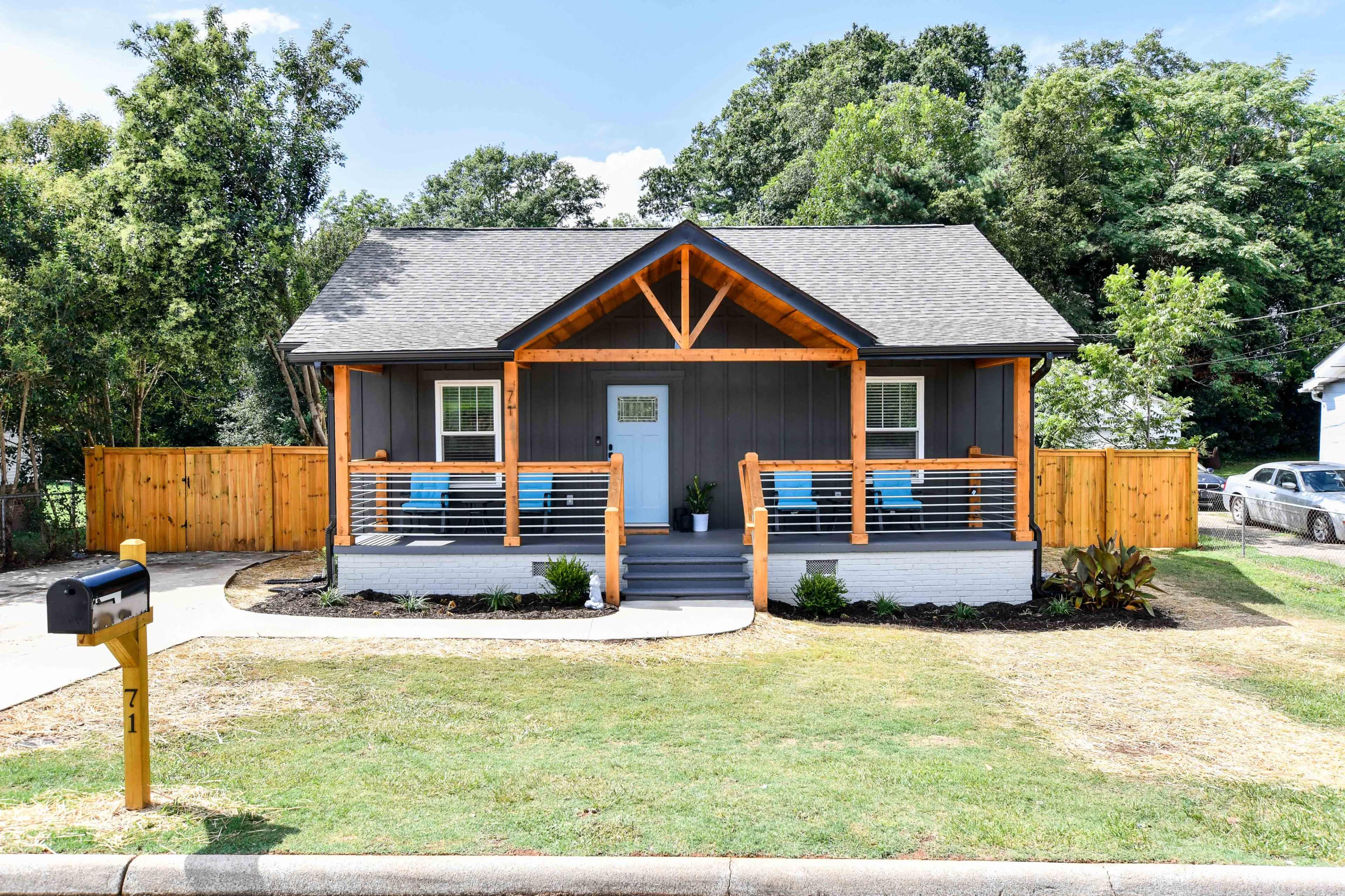 A single-story gray house with orange accents, a porch with blue chairs, and a wooden fence is set against a backdrop of trees.