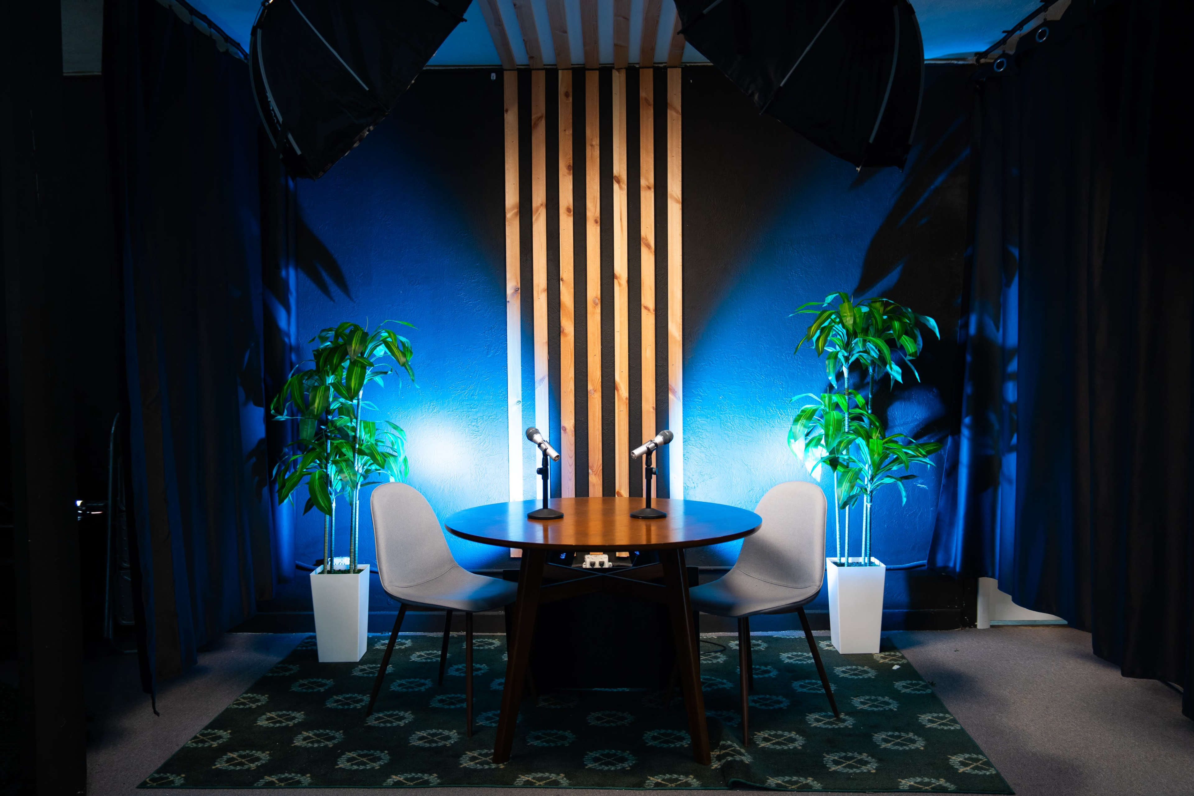 A small studio set-up features a round wooden table with two microphones and two chairs, surrounded by black curtains and illuminated by soft blue lighting with potted plants in white stands.