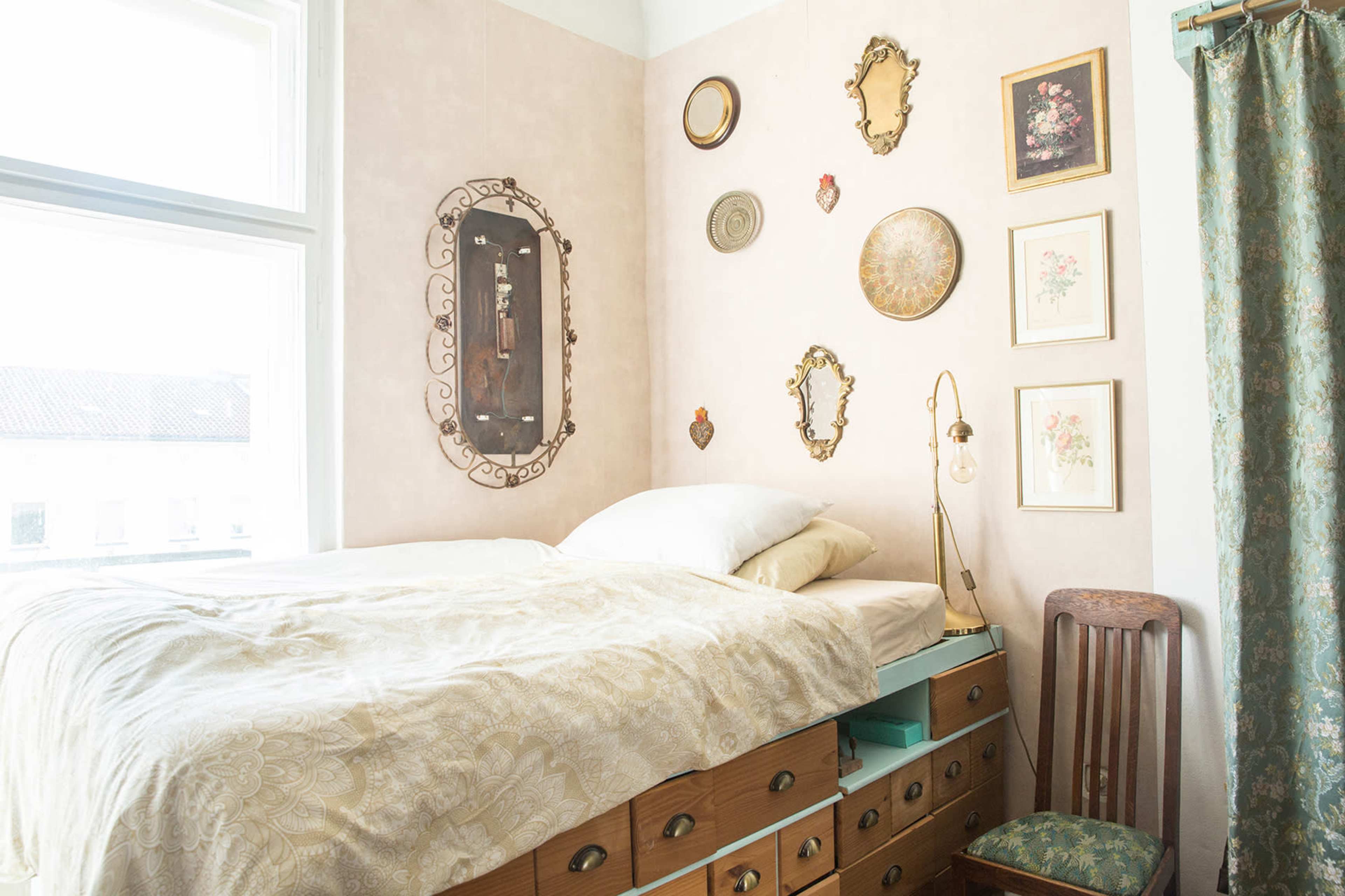 The image shows a cozy bedroom corner featuring a bed with a patterned blanket, surrounded by various framed artworks and decorative mirrors on the pale wall.