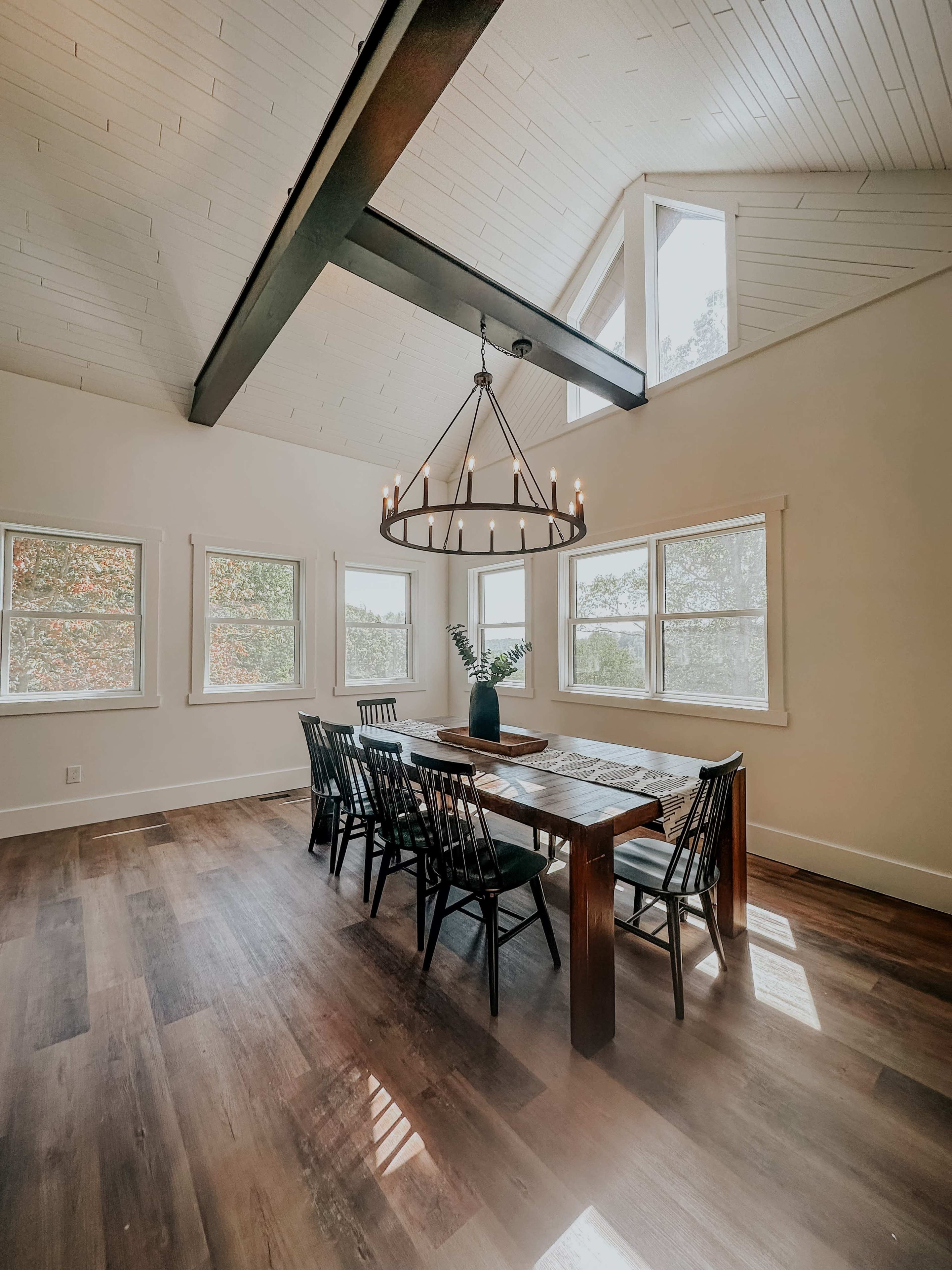 A spacious dining area features a large wooden table surrounded by black chairs beneath a chandelier, with several windows allowing natural light to fill the room.