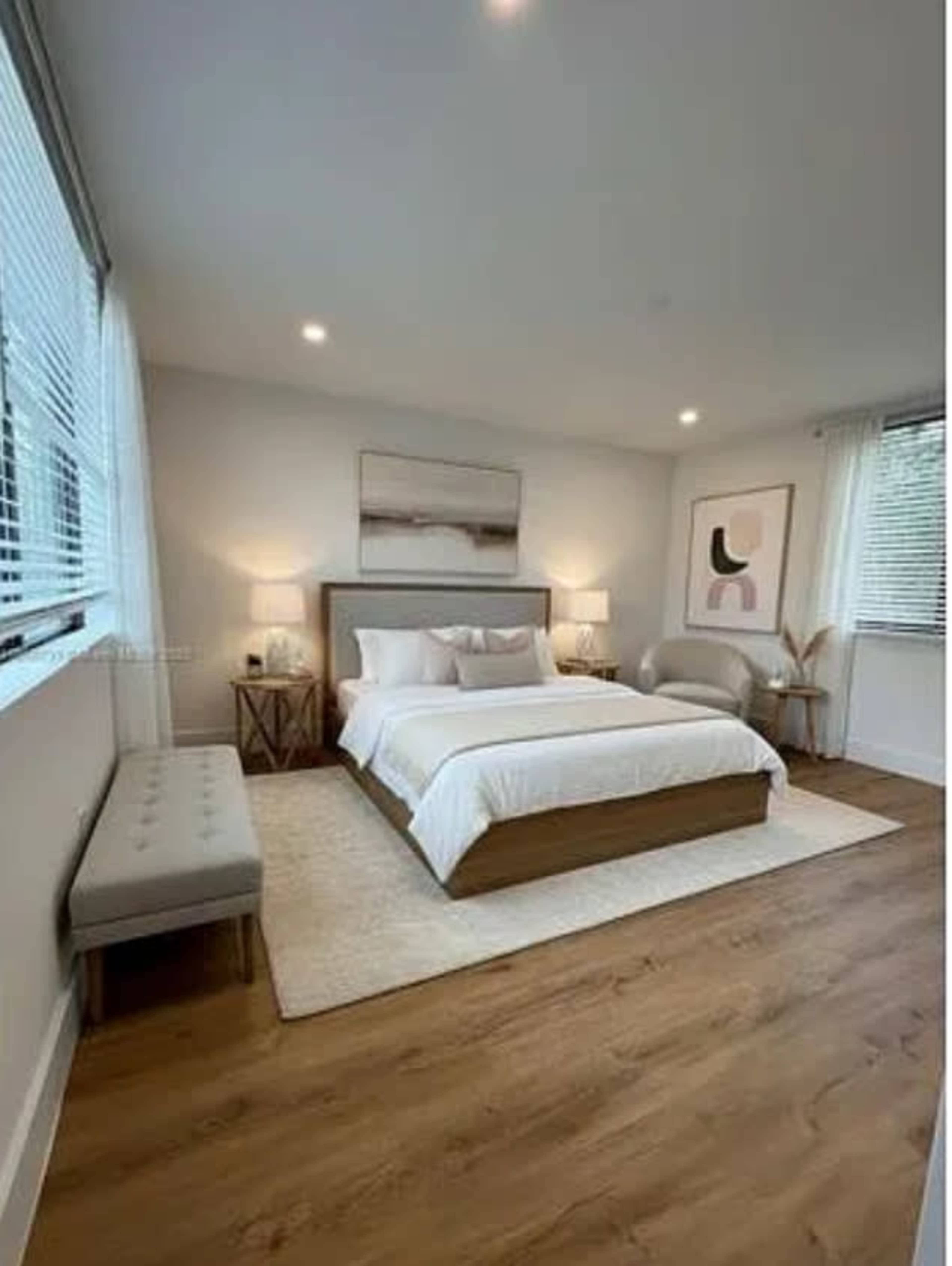 A modern bedroom features a large bed with white bedding, a gray headboard, and natural wood accents, along with a cozy seating area and large windows letting in light.