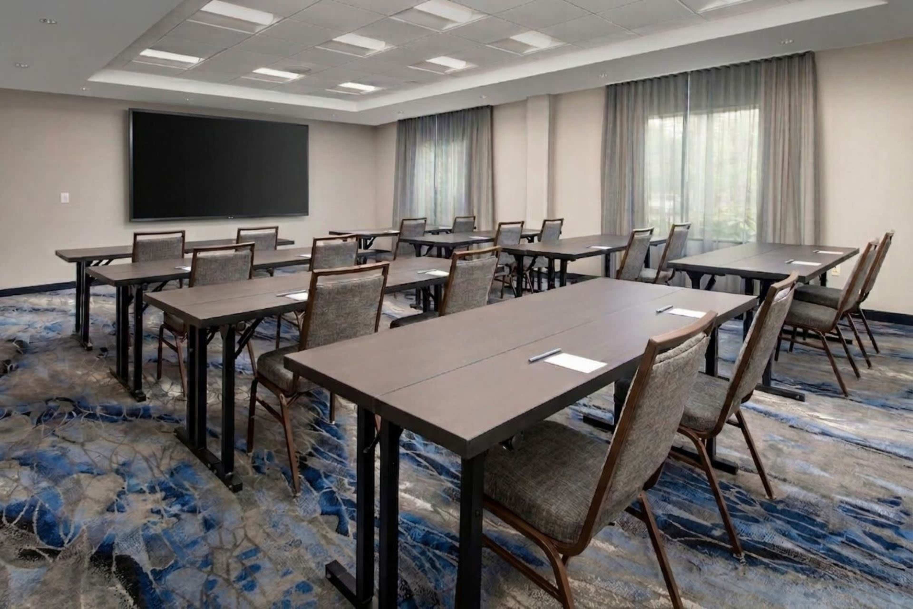 The image shows a conference room setup with several rectangular tables and chairs arranged in a classroom style, featuring a large screen at the front and windows with sheer curtains.