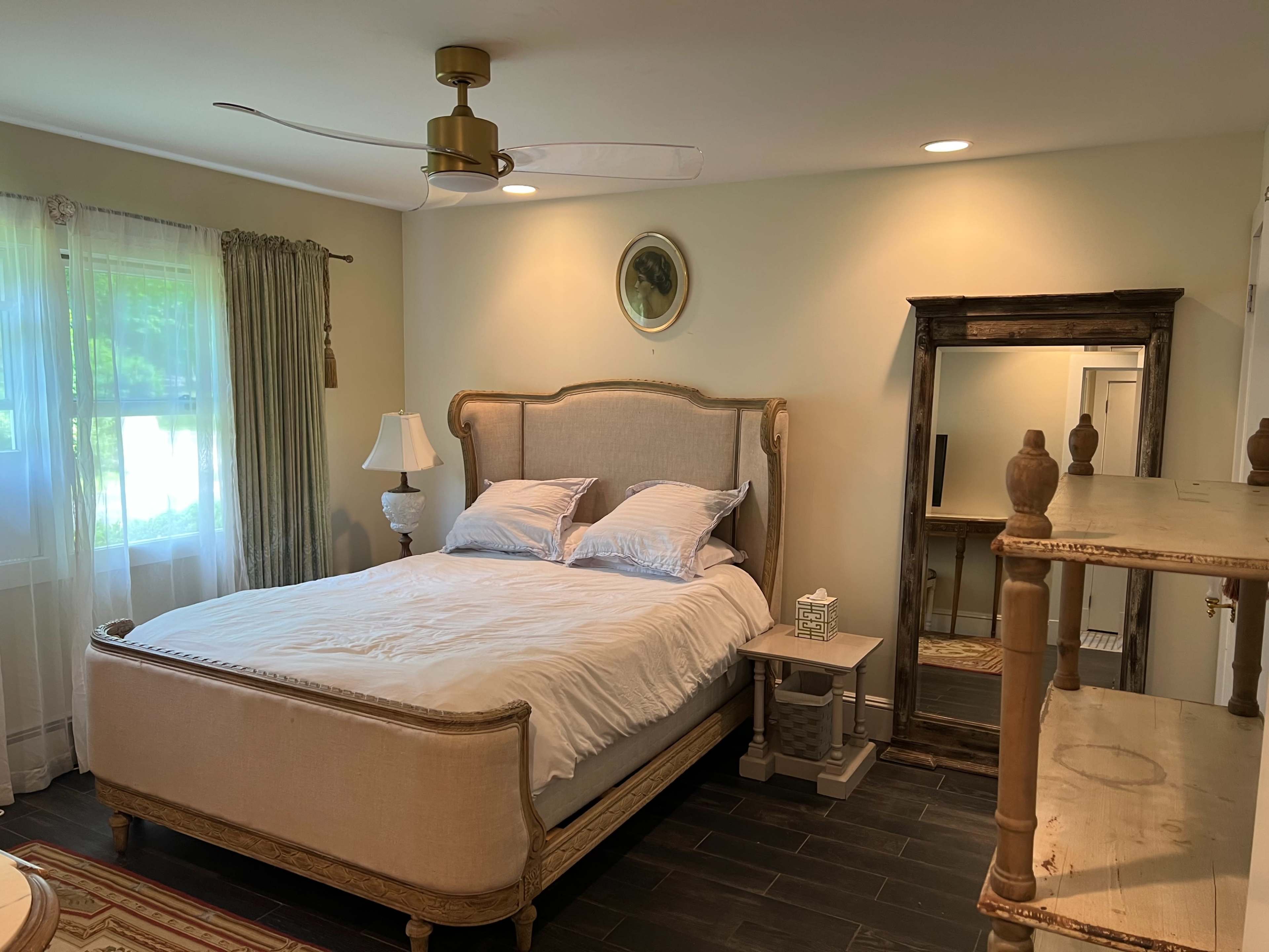 A bedroom with a large bed, a window with sheer curtains, and a mirror along with a bedside table and decorative shelf.