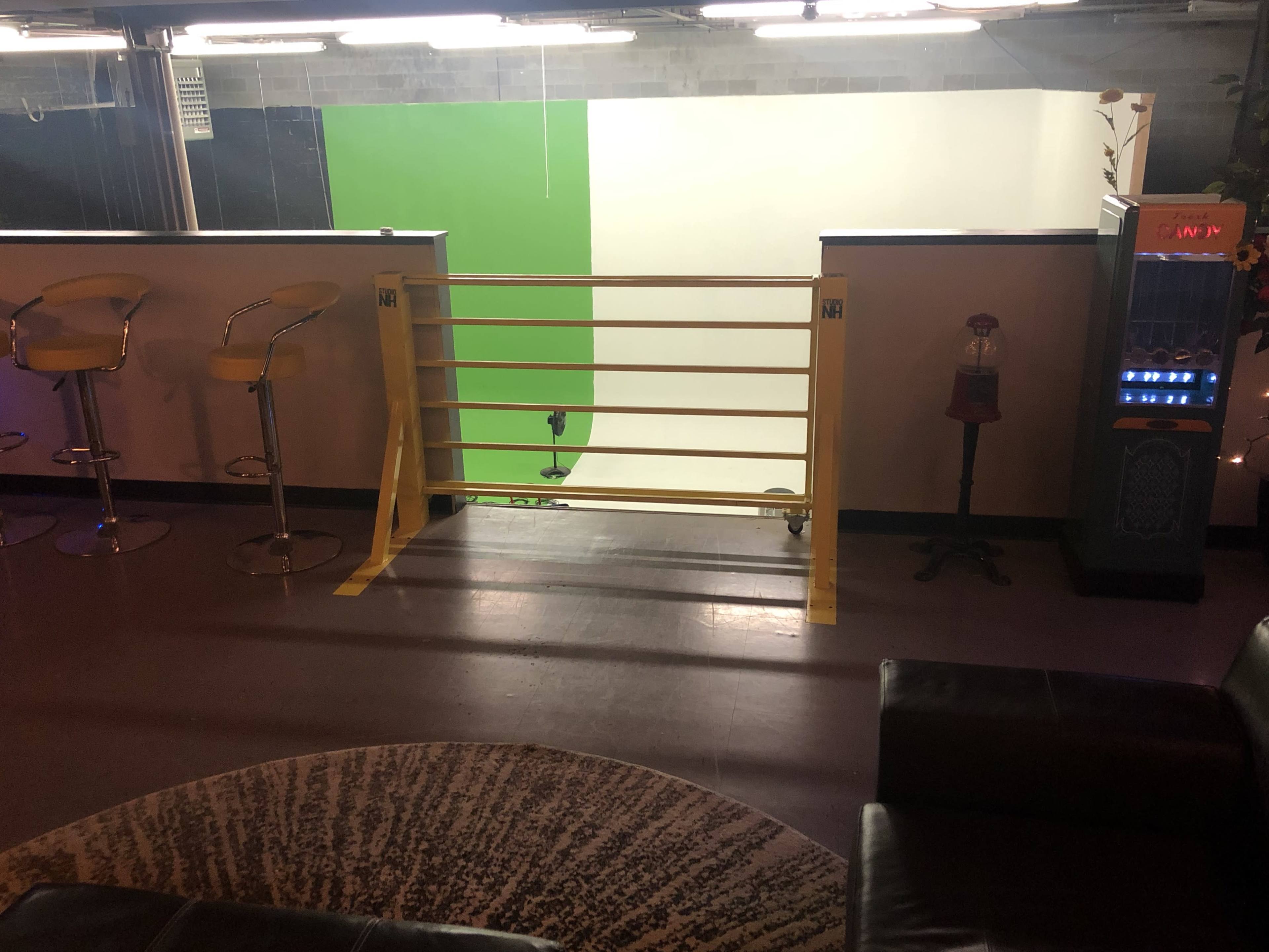 A room with a railing overlooking a green and white studio backdrop, a sitting area with a rug and couch, and a vending machine nearby.