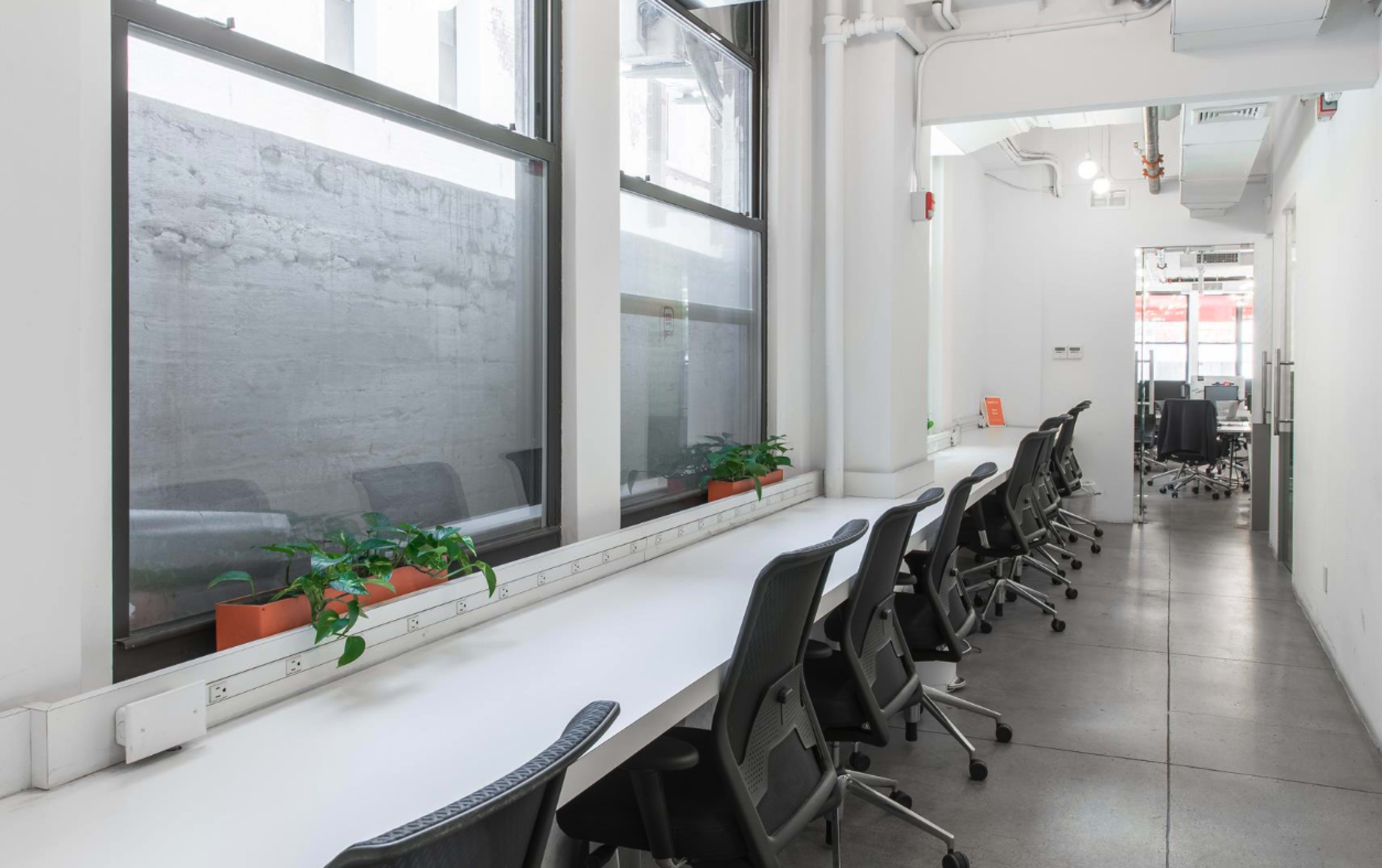The image shows a modern office space with a row of empty chairs along a long white desk, flanked by large windows with concrete walls visible outside.