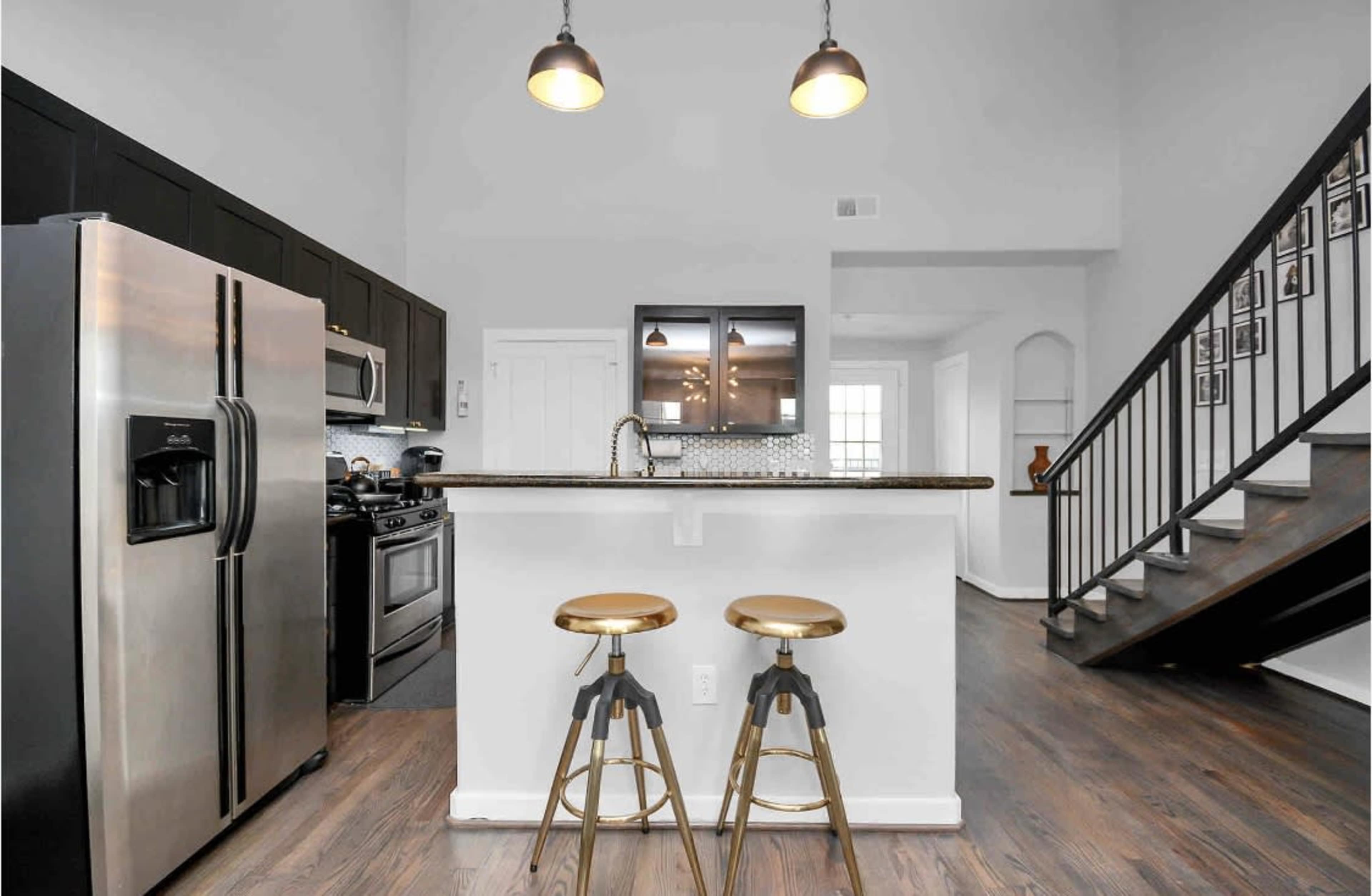 A modern kitchen features black cabinetry, stainless steel appliances, a central island with two stools, and a staircase in the background.