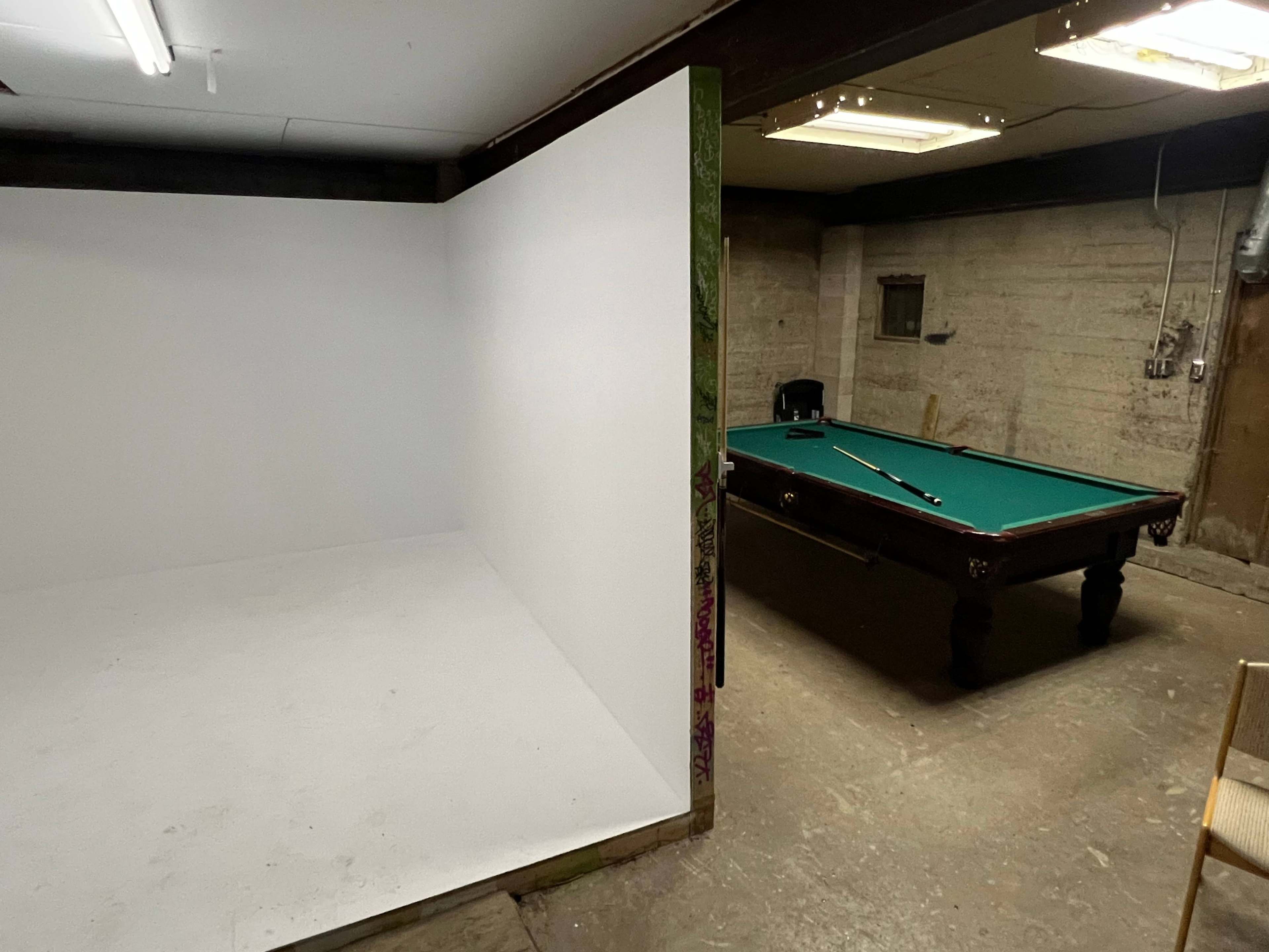 A bare white room is located adjacent to a pool table in a dimly lit basement.