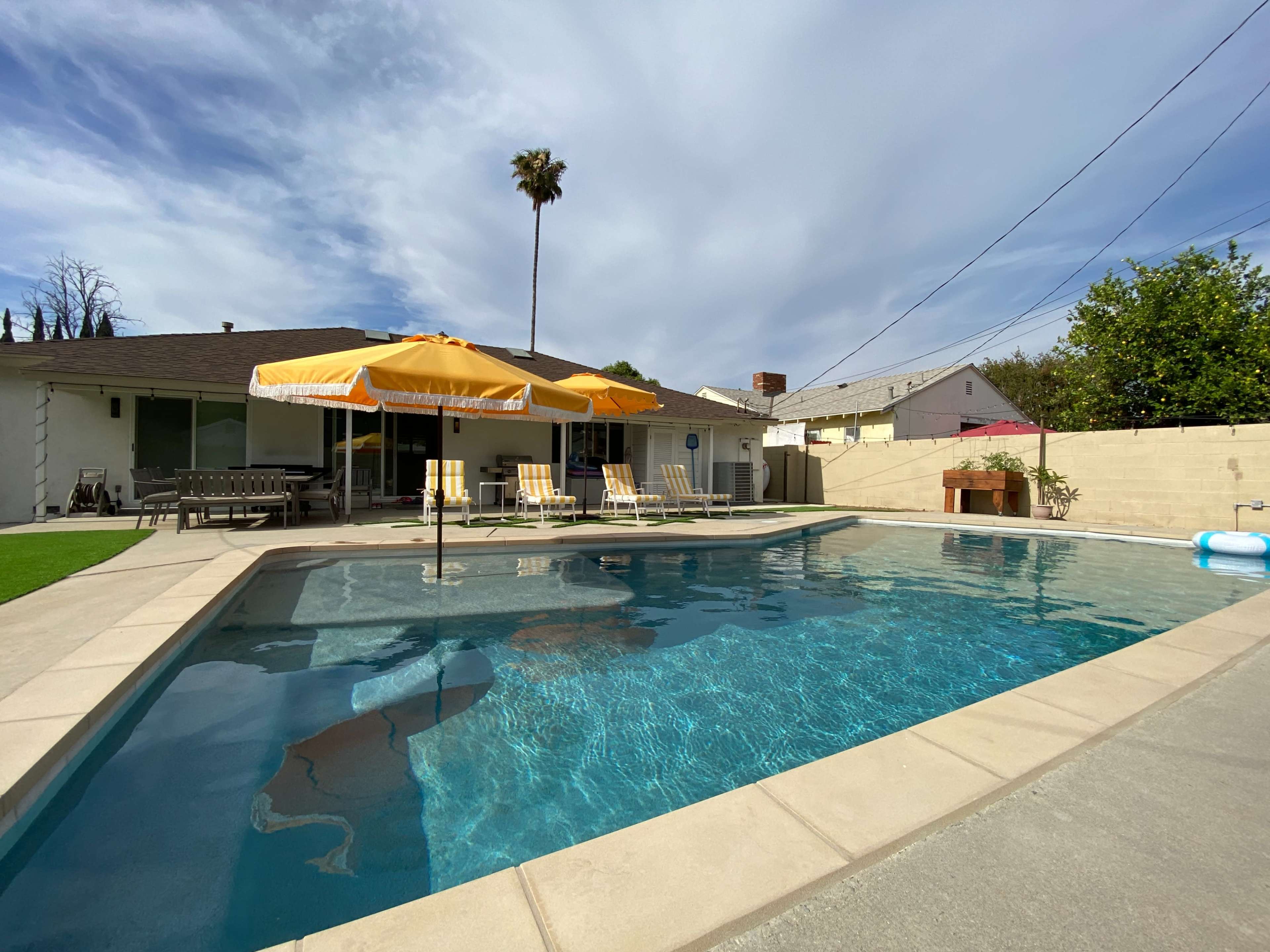 A rectangular swimming pool surrounded by lounge chairs and two yellow umbrellas beside a single-story house with a palm tree in the background.