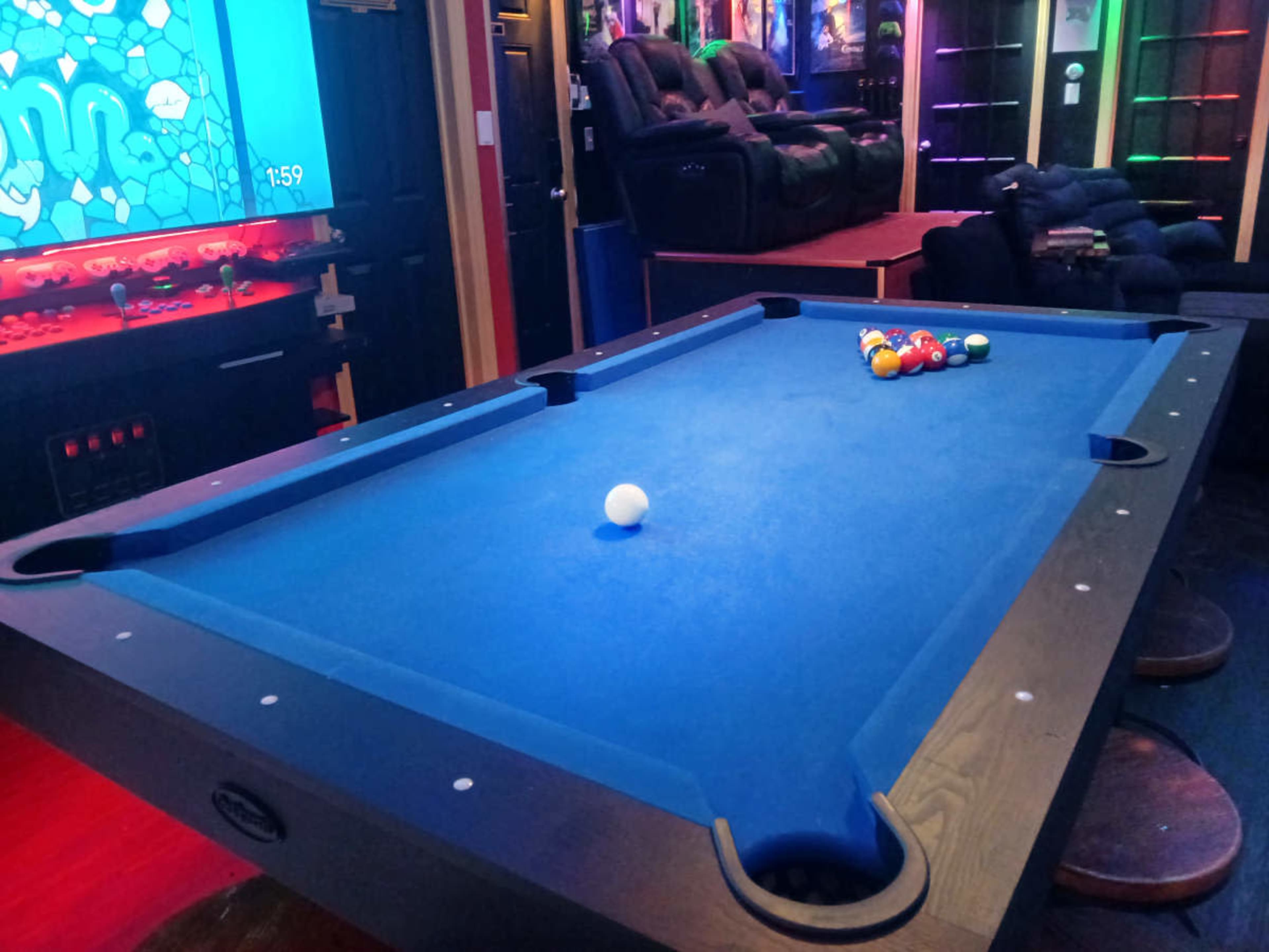 A blue pool table with a white cue ball and a rack of colorful billiard balls is positioned in a game room with dim lighting and a couch in the background.