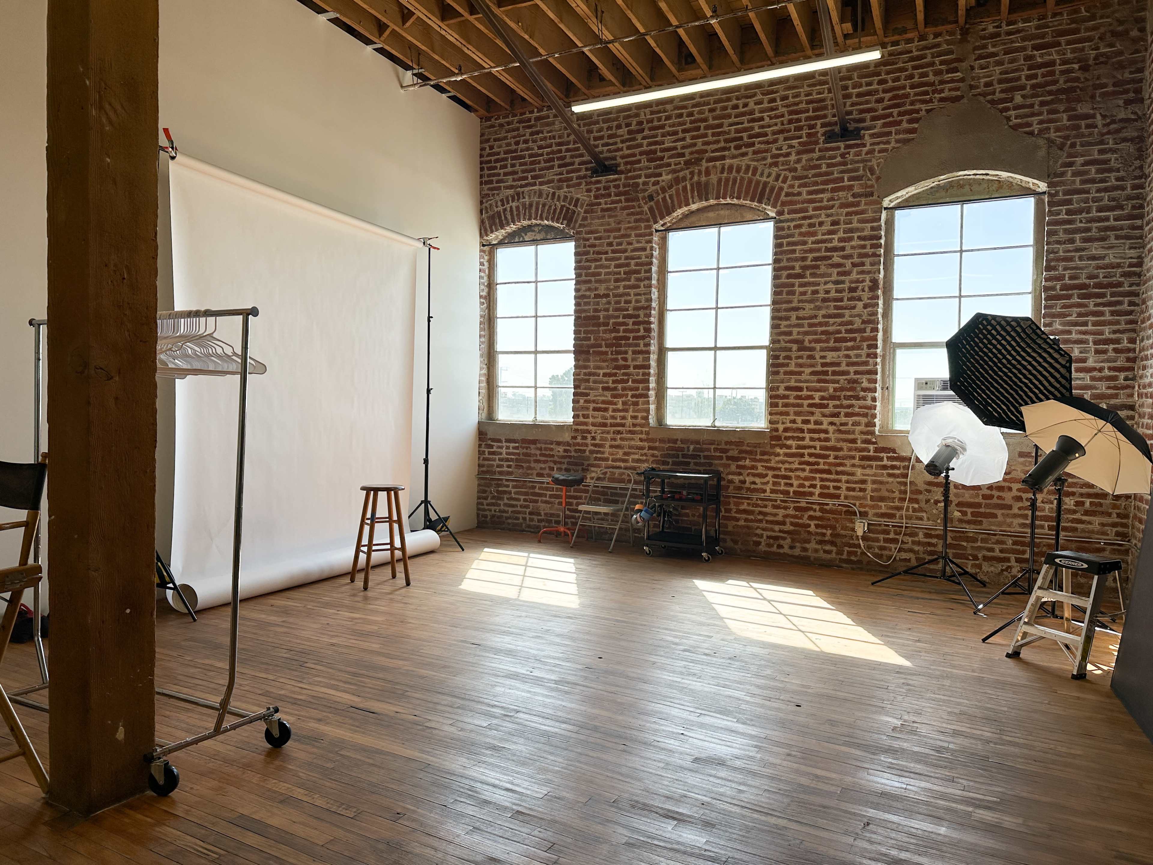 The image depicts a photography studio featuring brick walls, large windows, a wooden floor, and various photography equipment.