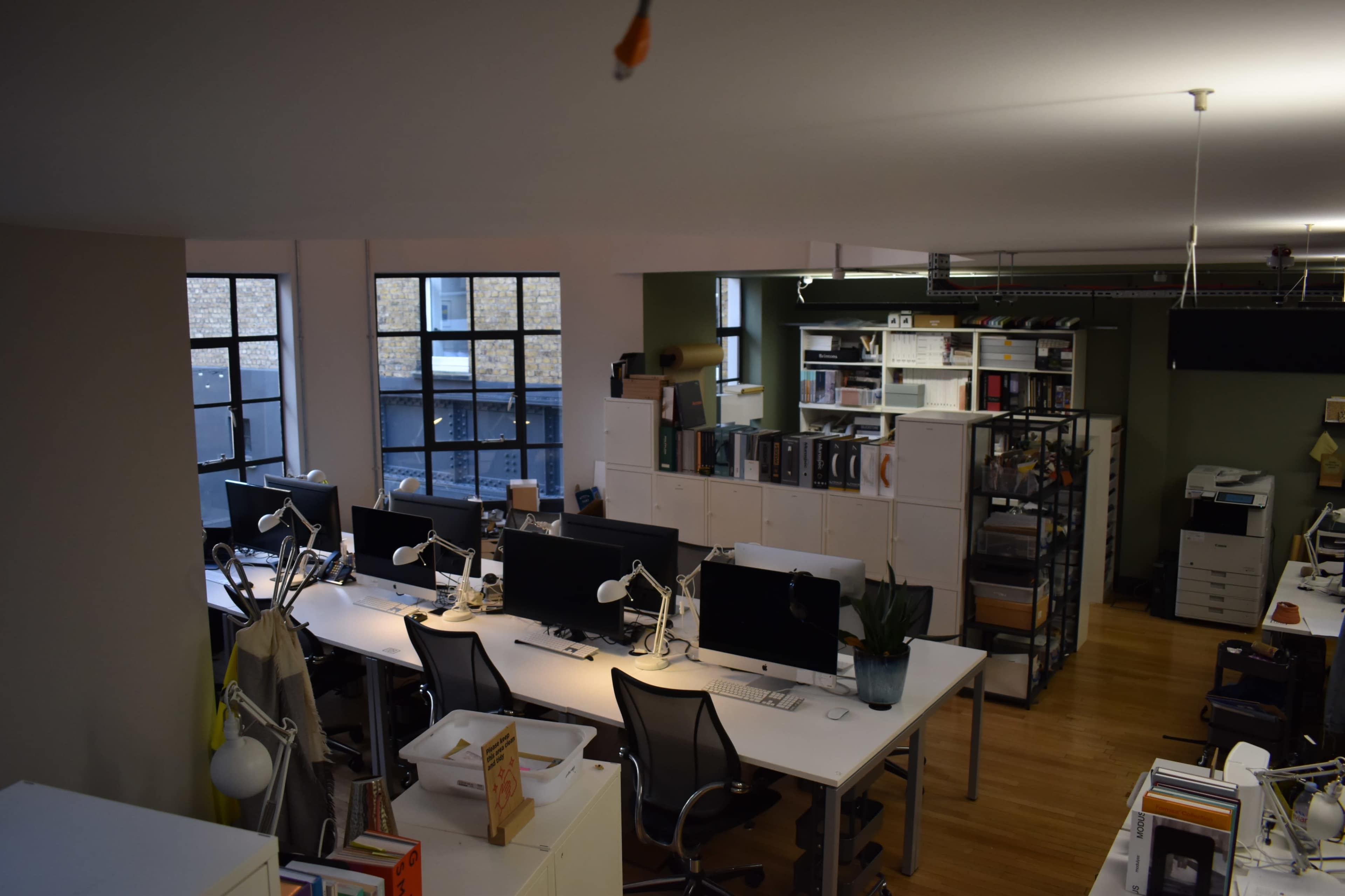 The image shows an open office space with several desks equipped with computers and shelves filled with documents and supplies.