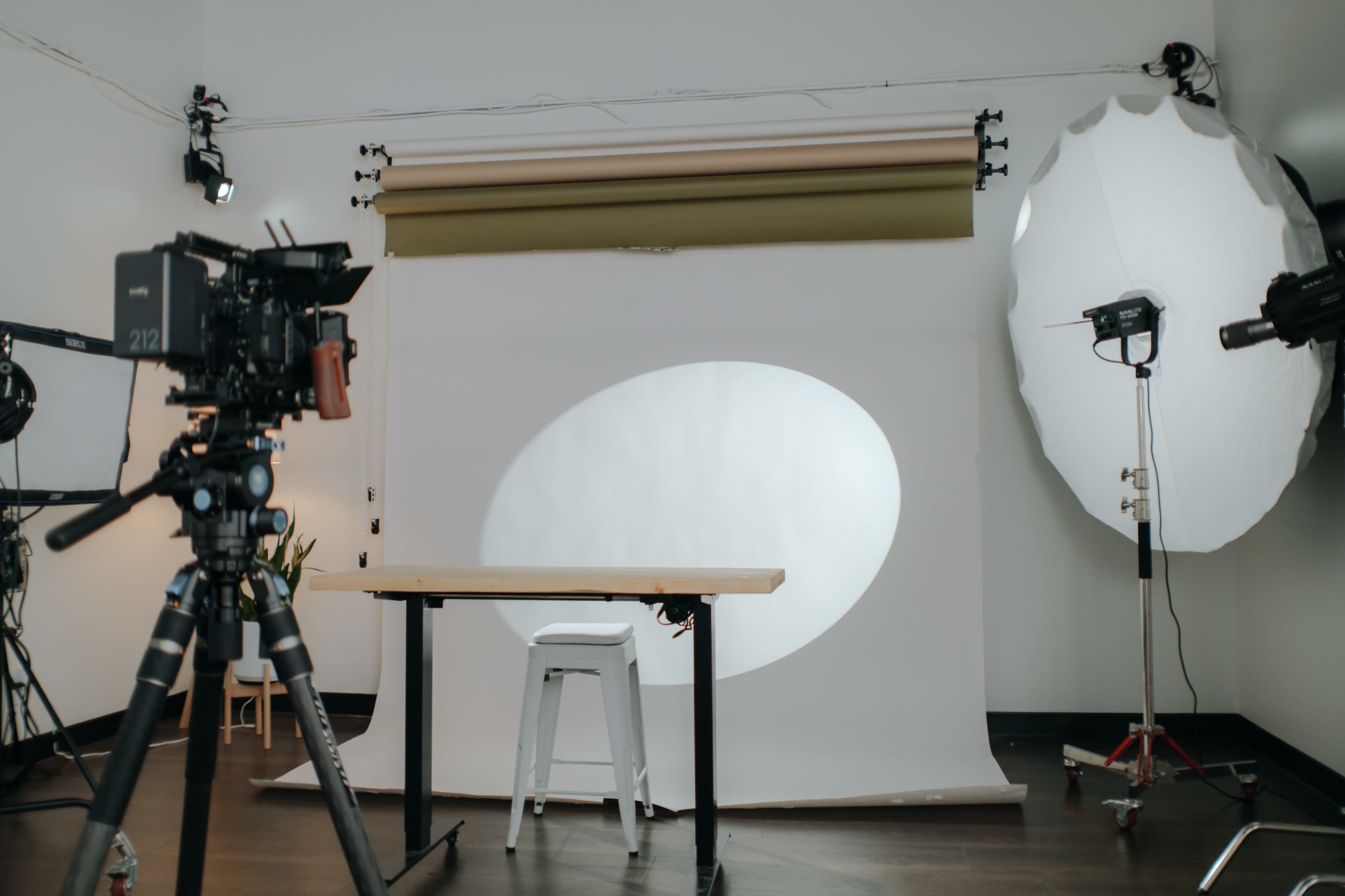 A photography studio setup features a backdrop with a light spot, a table, a stool, and lighting equipment.