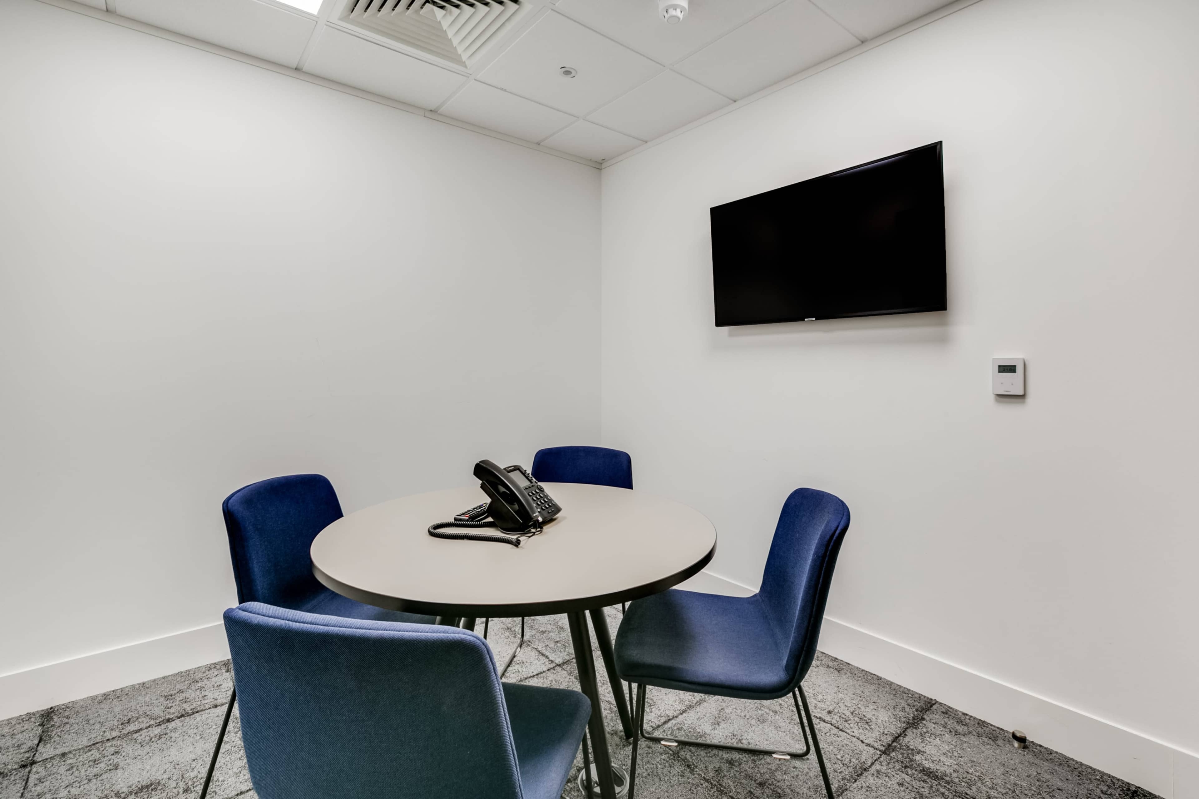 A small meeting room features a round table with four blue chairs, a telephone, and a wall-mounted television.