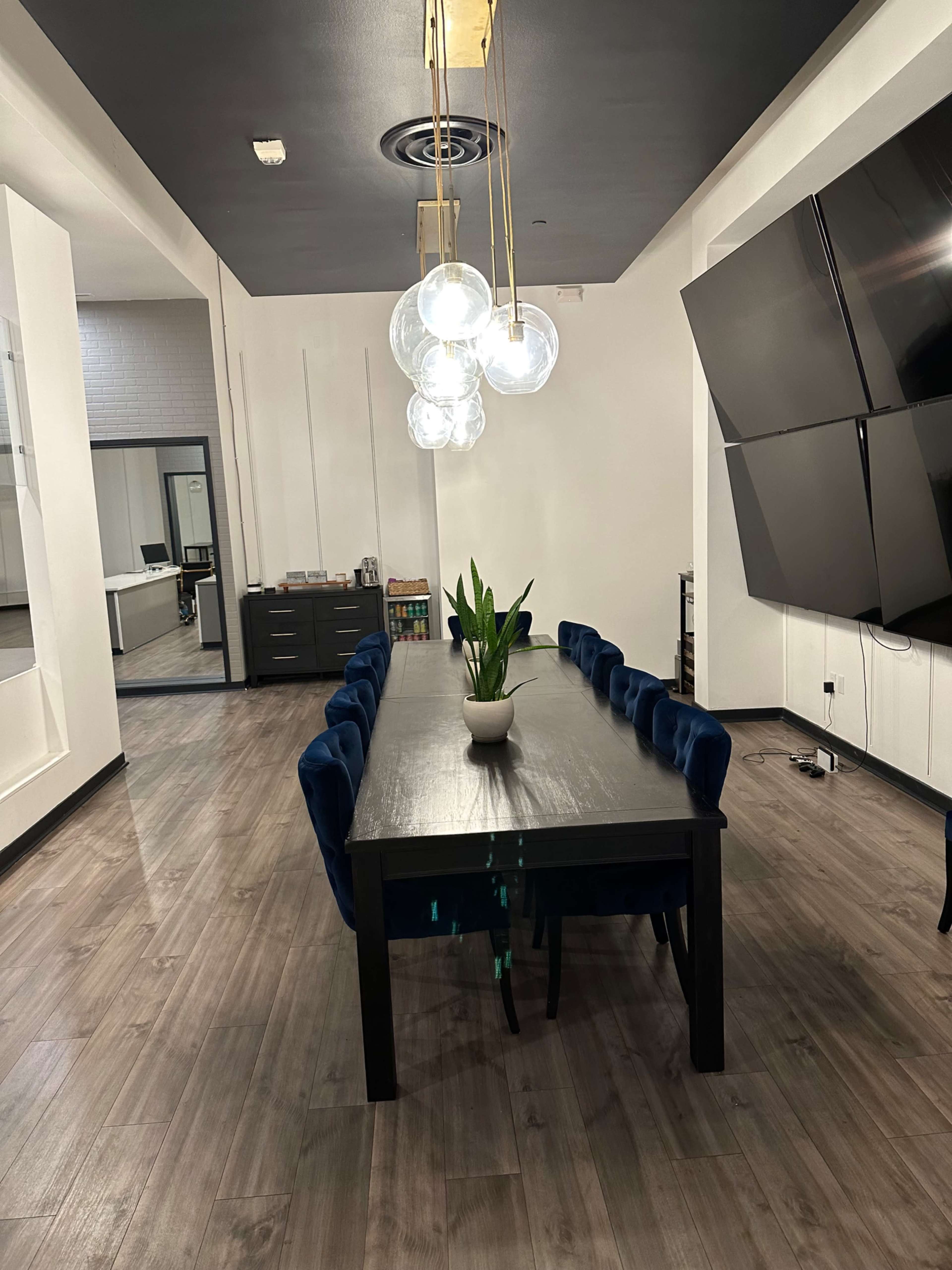 A spacious dining area with a long black table surrounded by blue chairs, illuminated by hanging light fixtures, and a potted plant at the center.