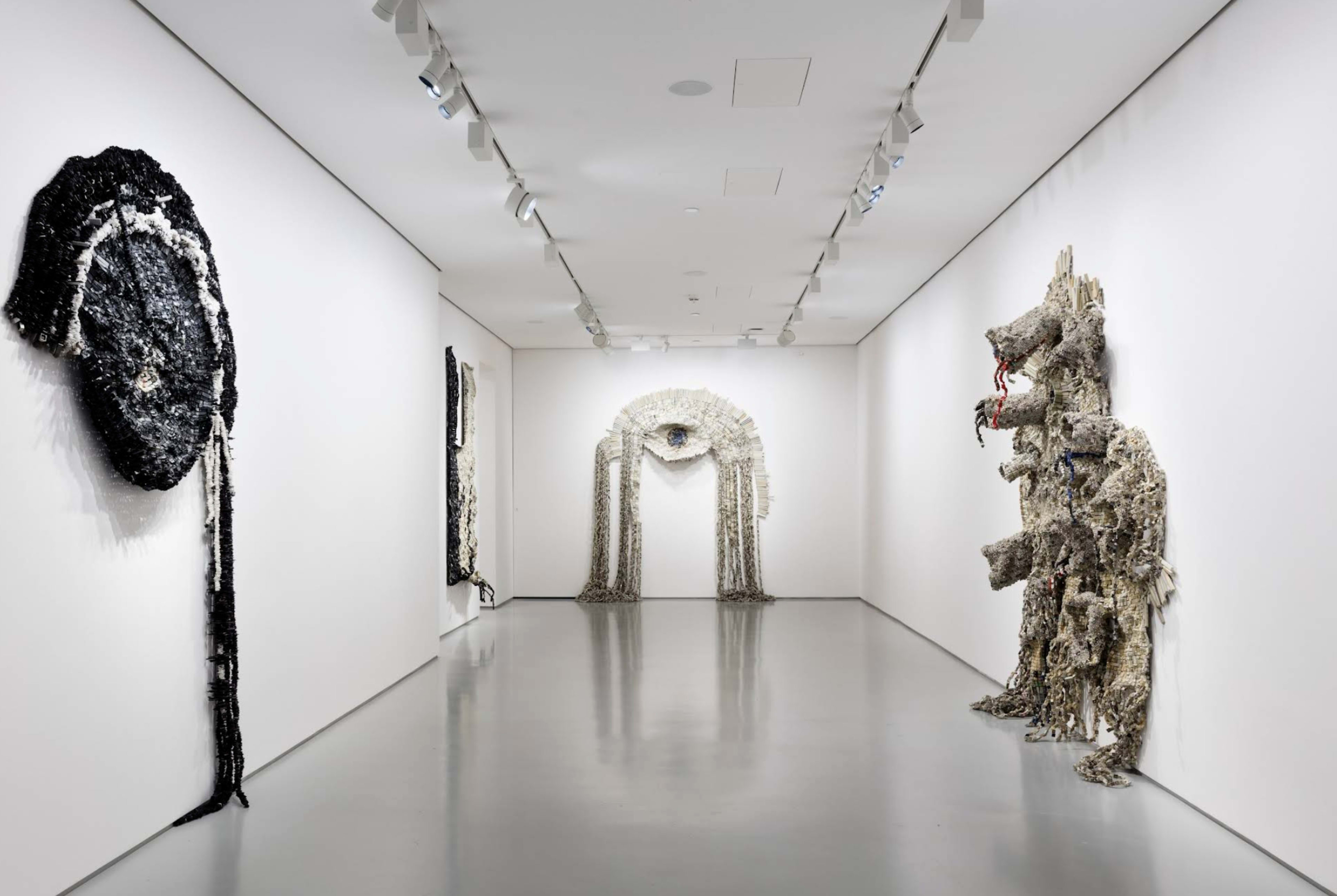The image shows an art gallery with textured wall hangings on display, including an arched piece and a large figure resembling a creature, all set against plain white walls and a polished floor.