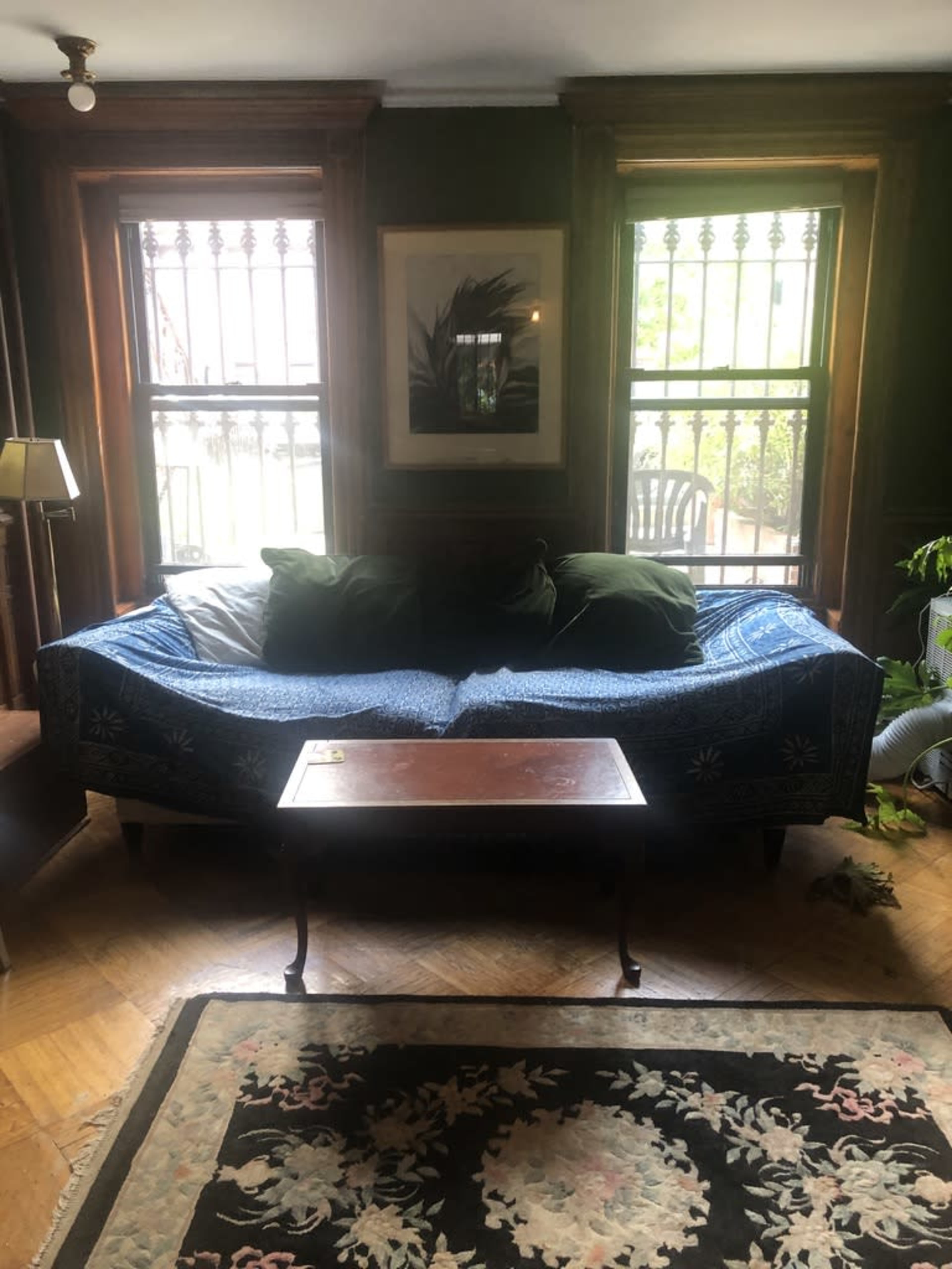 A blue-patterned couch with green throw pillows sits in front of a coffee table in a room featuring large windows and a patterned rug.