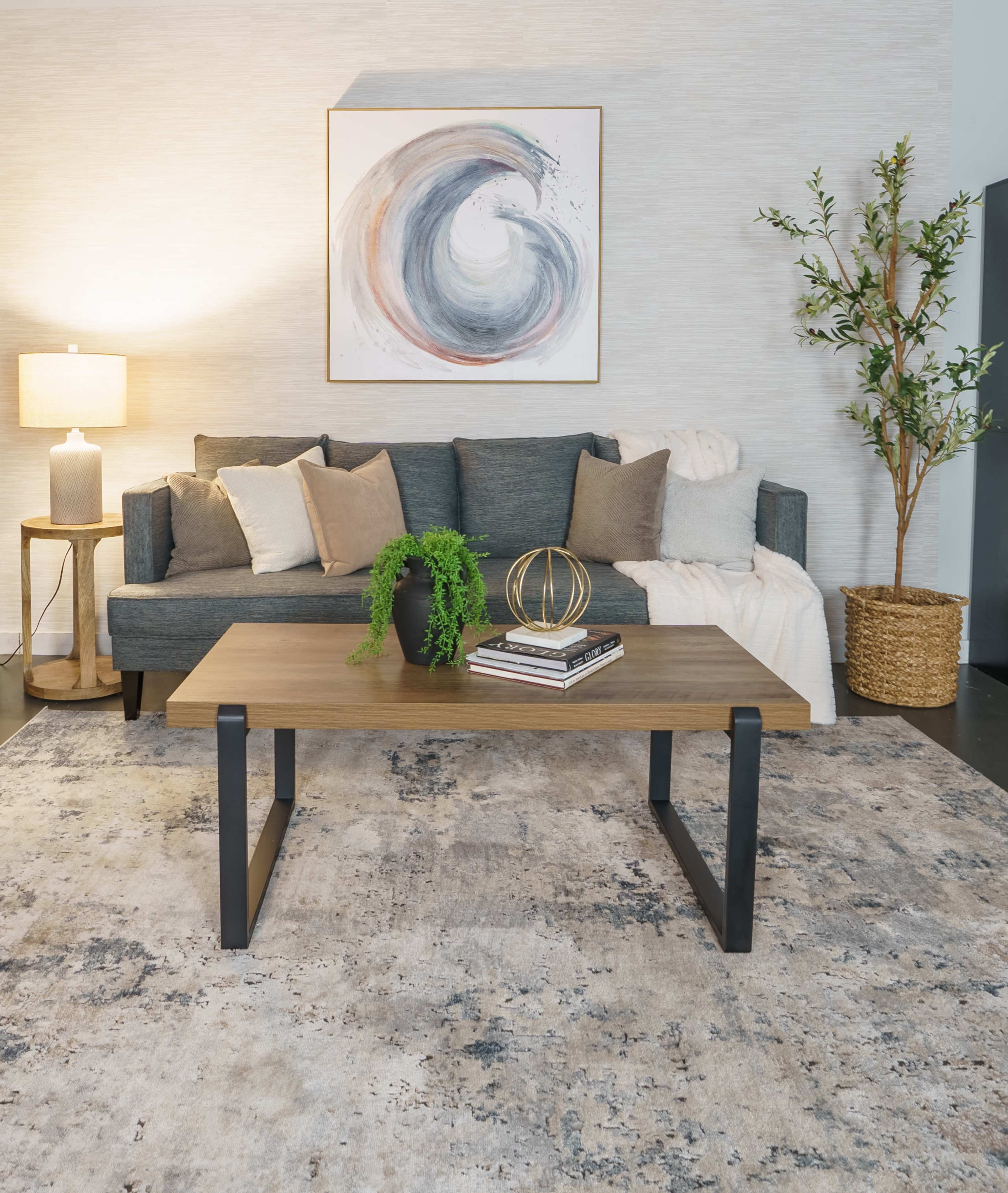 A cozy living room with a gray sofa, a wooden coffee table, a round lamp, decorative pillows, a potted plant, and a wall-mounted artwork featuring a swirling design.